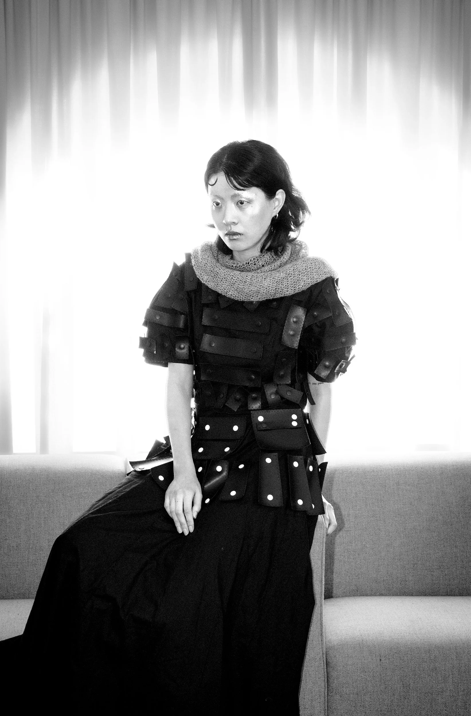 A woman with short dark hair sitting on a bench in front of a window with closed curtains, wearing a black dress with a polka dot skirt and a knitted scarf around her neck.