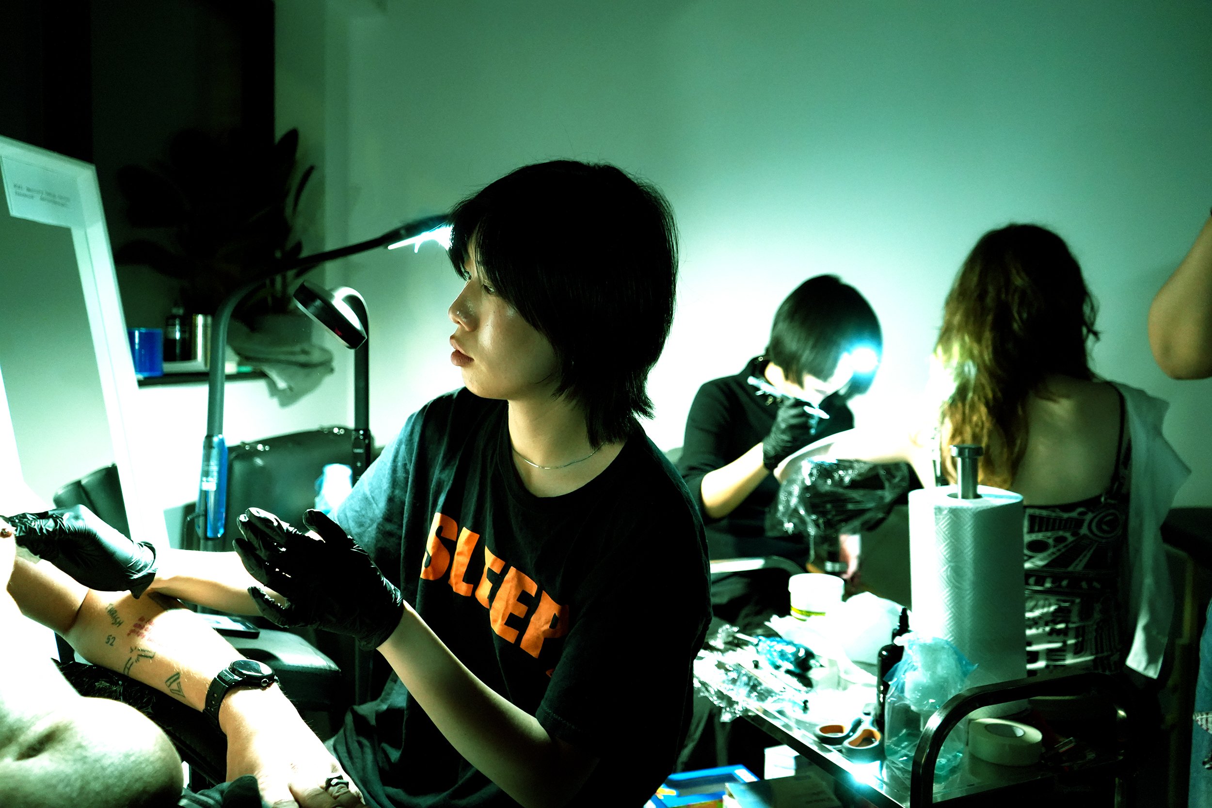 A woman with short black hair getting a tattoo, while another person works on a tattoo in the background. The scene takes place in a dimly lit room with a greenish hue.