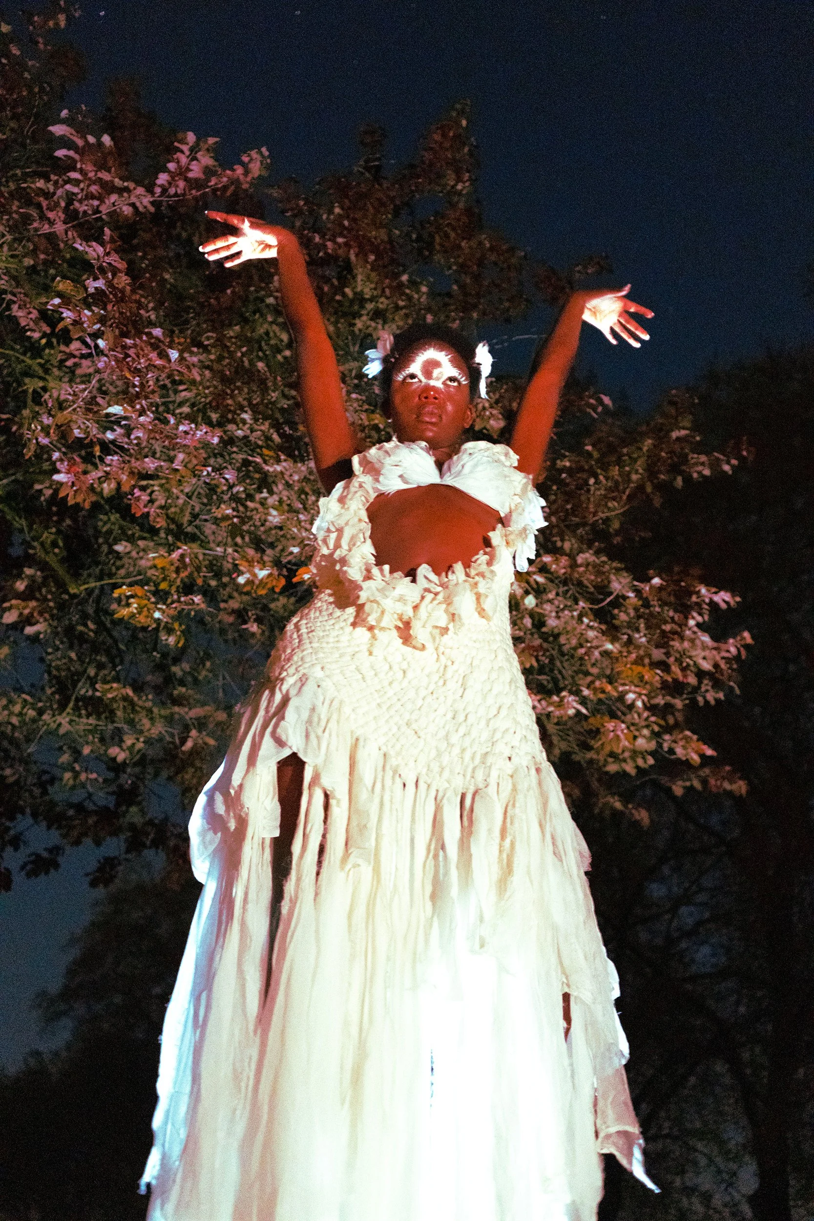 A person dressed in a white, textured, flowy dress with ruffles, standing outdoors at night, with their arms raised. They have face paint around their eyes and face and are wearing flowers in their hair. Behind them are trees and a dark night sky.