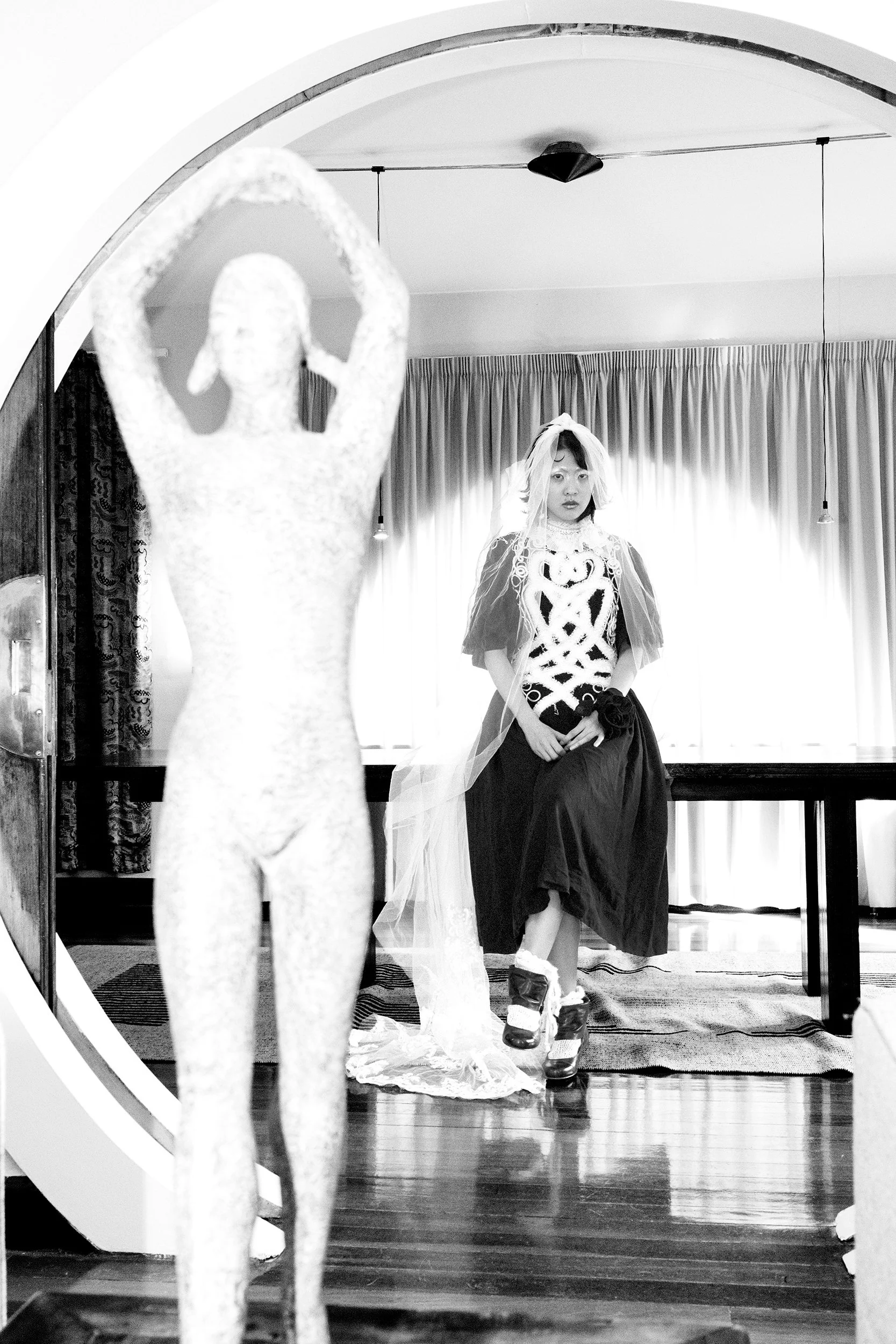 A woman dressed in vintage clothing, including a veil, sitting indoors in front of curtains, with a mannequin in the foreground.