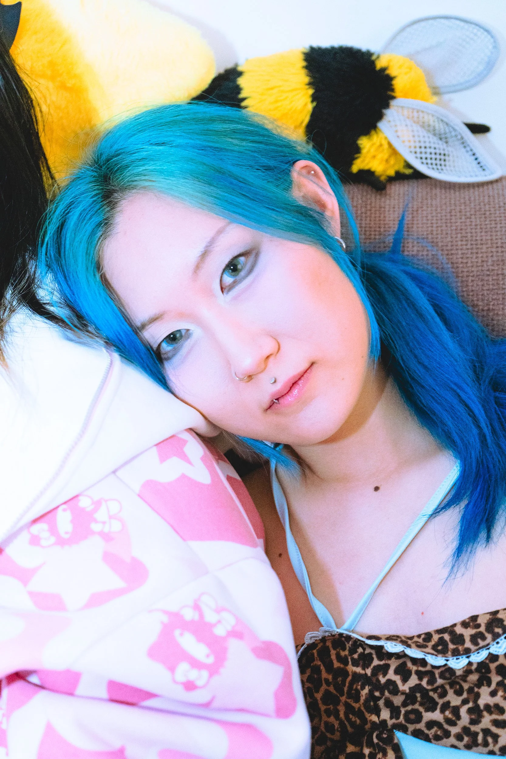 A woman with blue hair lying on a bed next to a pillow and wall decoration, wearing a leopard print dress