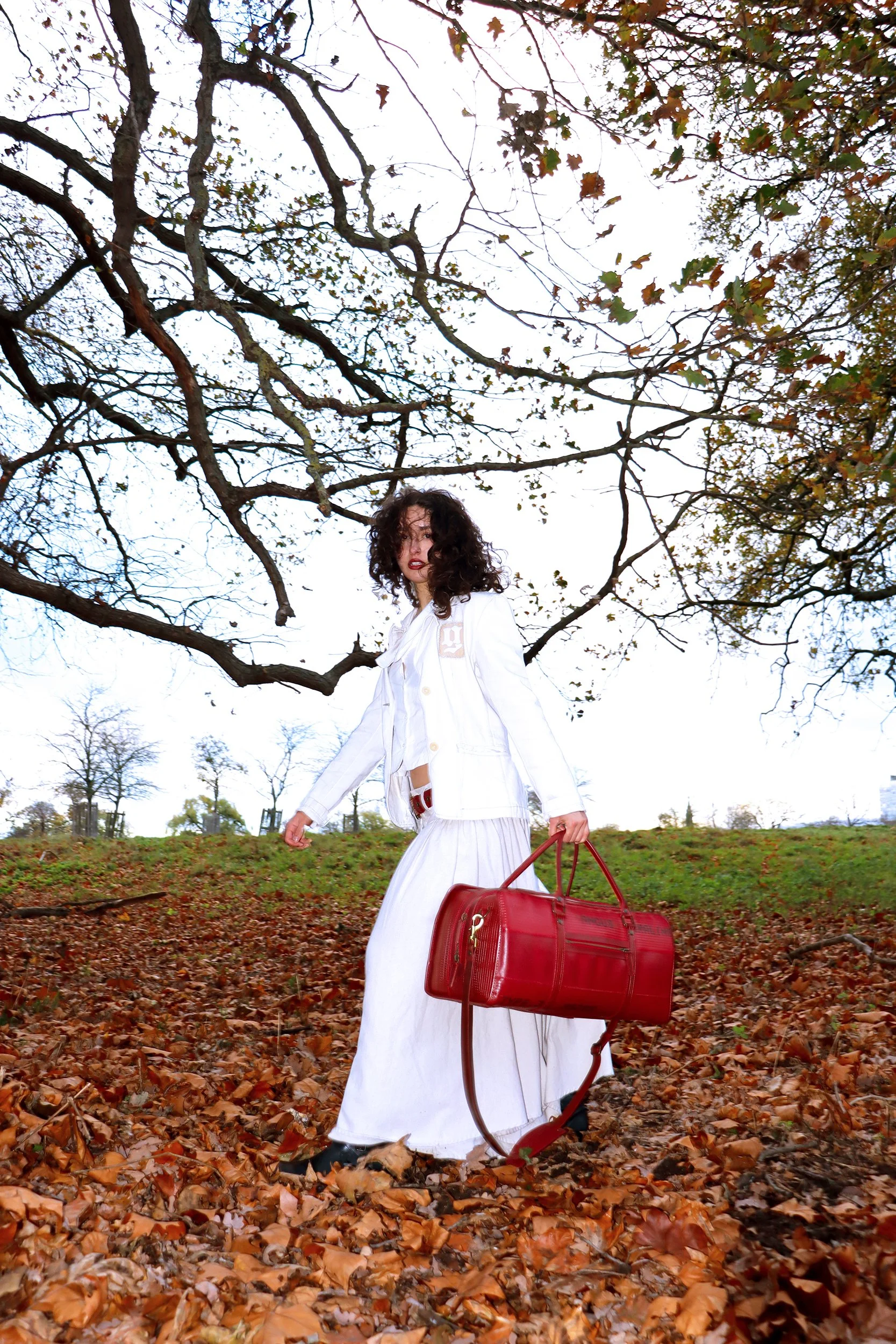 A woman with curly dark hair wearing a white outfit, holding a red bag, walking through a park with fallen leaves, trees with mostly bare branches, and a bright sky in the background.