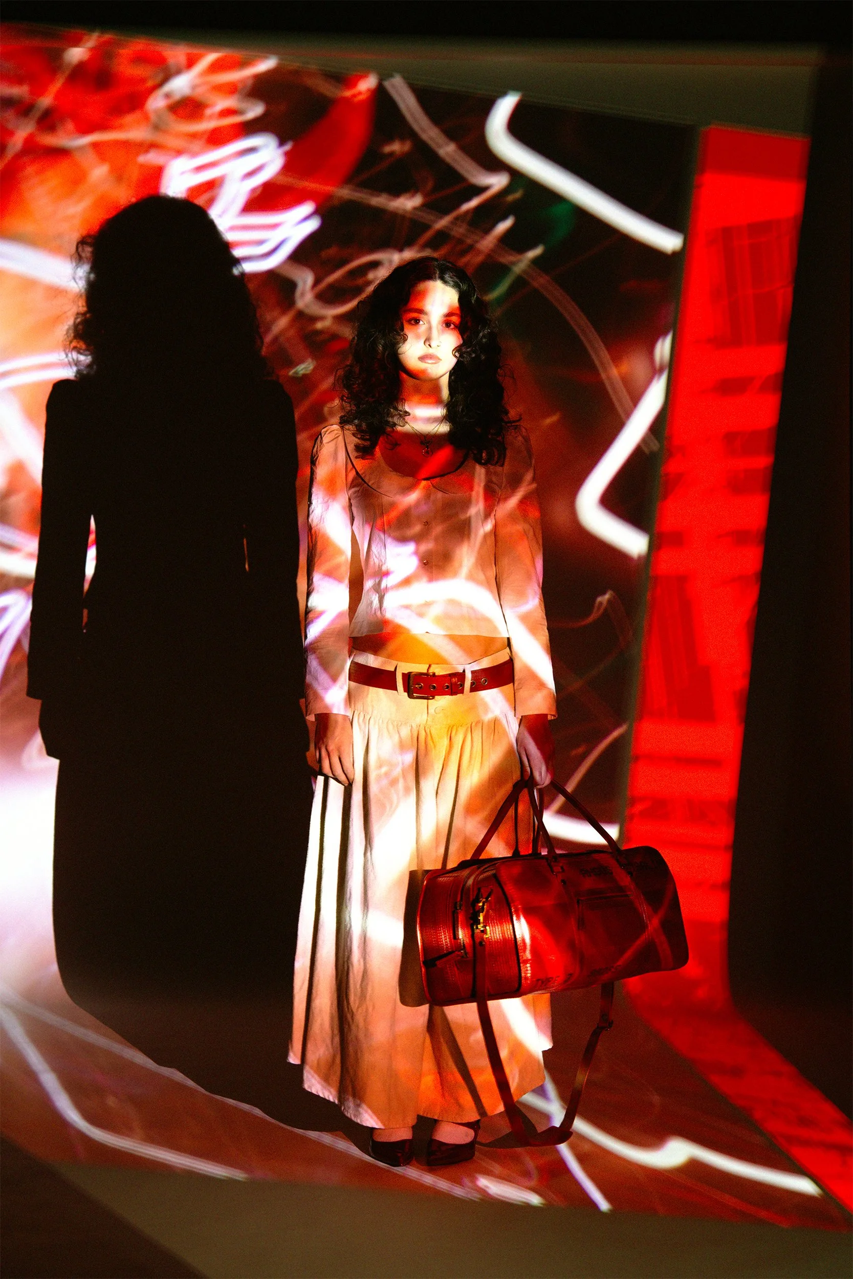 Young woman with curly dark hair wearing a silk floral blouse and long skirt, standing in front of a colorful, abstract light display with her shadow cast on the wall behind her. She holds a large red designer bag.