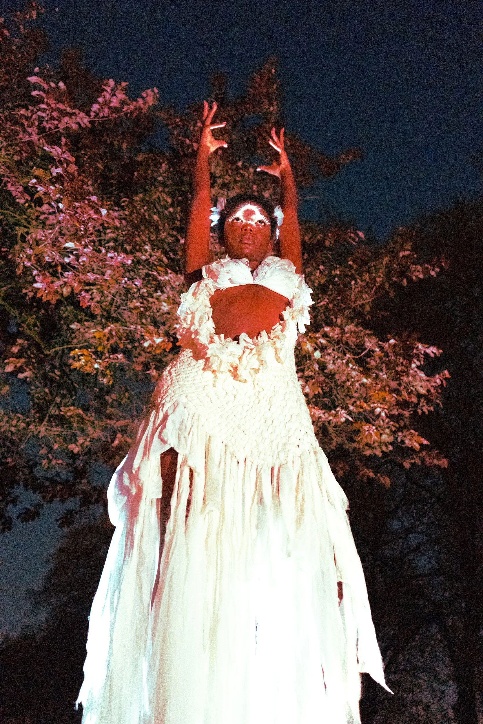A woman with face paint and floral accessories in a white dress, raising her hands with fingers forming 'peace' signs, standing outdoors at night against a dark sky and trees.