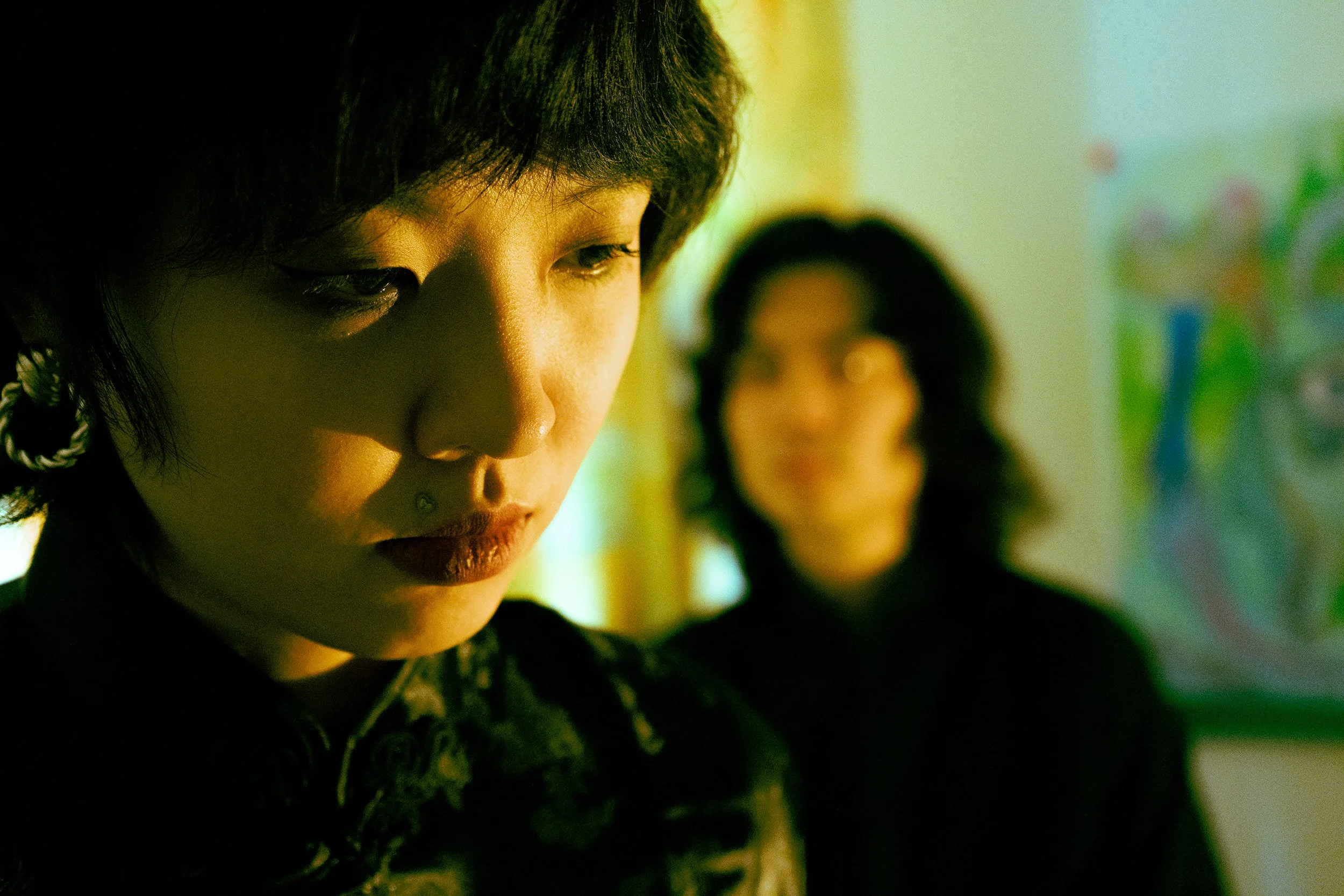 Close-up of a woman with short dark hair, looking down, with another woman blurred in the background. The lighting is moody and dramatic.