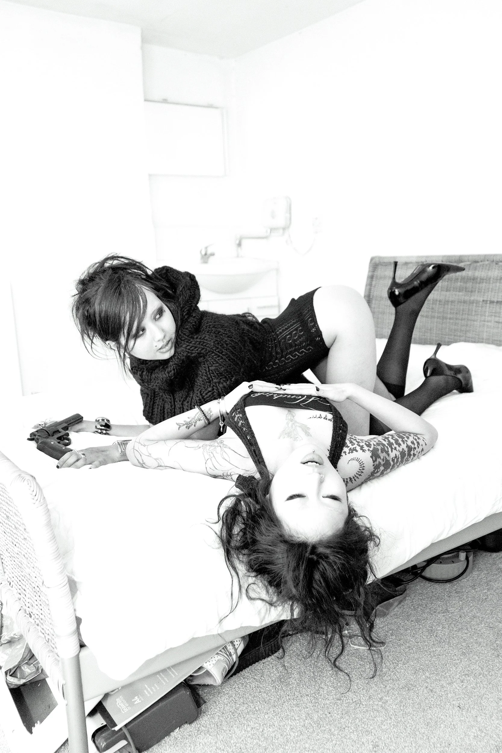 Four women are lying on a bed in a room with white walls. Two women are partially covered in black clothing and stockings, one woman has tattoos on her arm, and the fourth woman is lying with her eyes closed. There are guns and magazines on the bed near them.