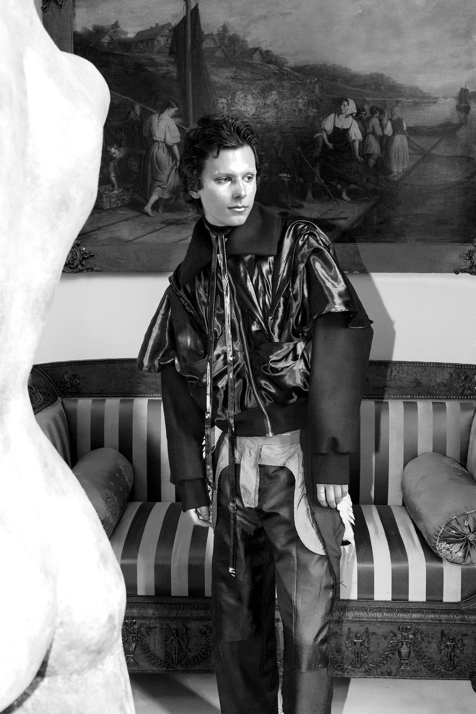 A young person with short dark hair wearing a shiny black jacket and baggy pants, standing in front of a painting of women by a river.