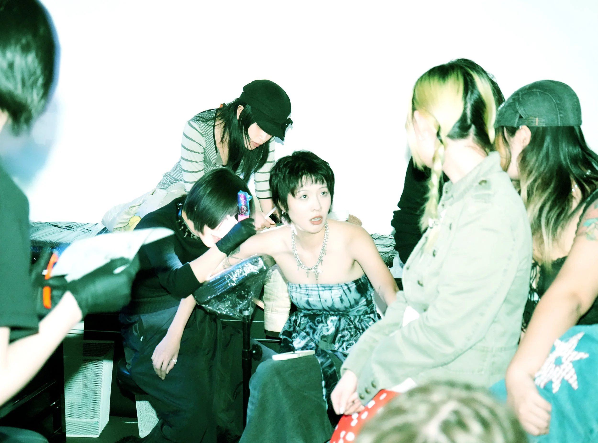 A group of people gathered around a woman with short dark hair, who is sitting shirtless and wearing a pearl necklace, as others look on and take photos.