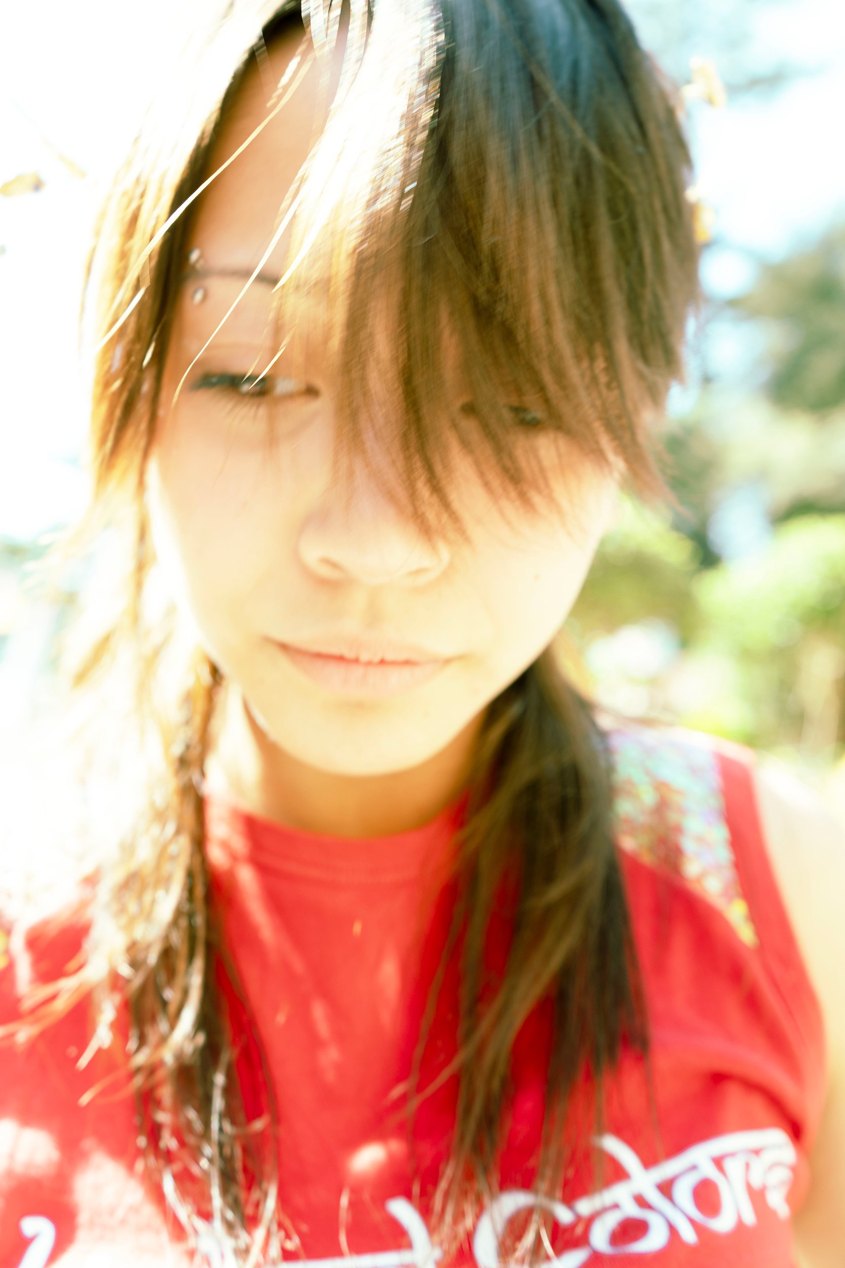 A close-up of a woman with long brown hair and bangs, wearing a red shirt with white text, outdoors with blurred greenery in the background.