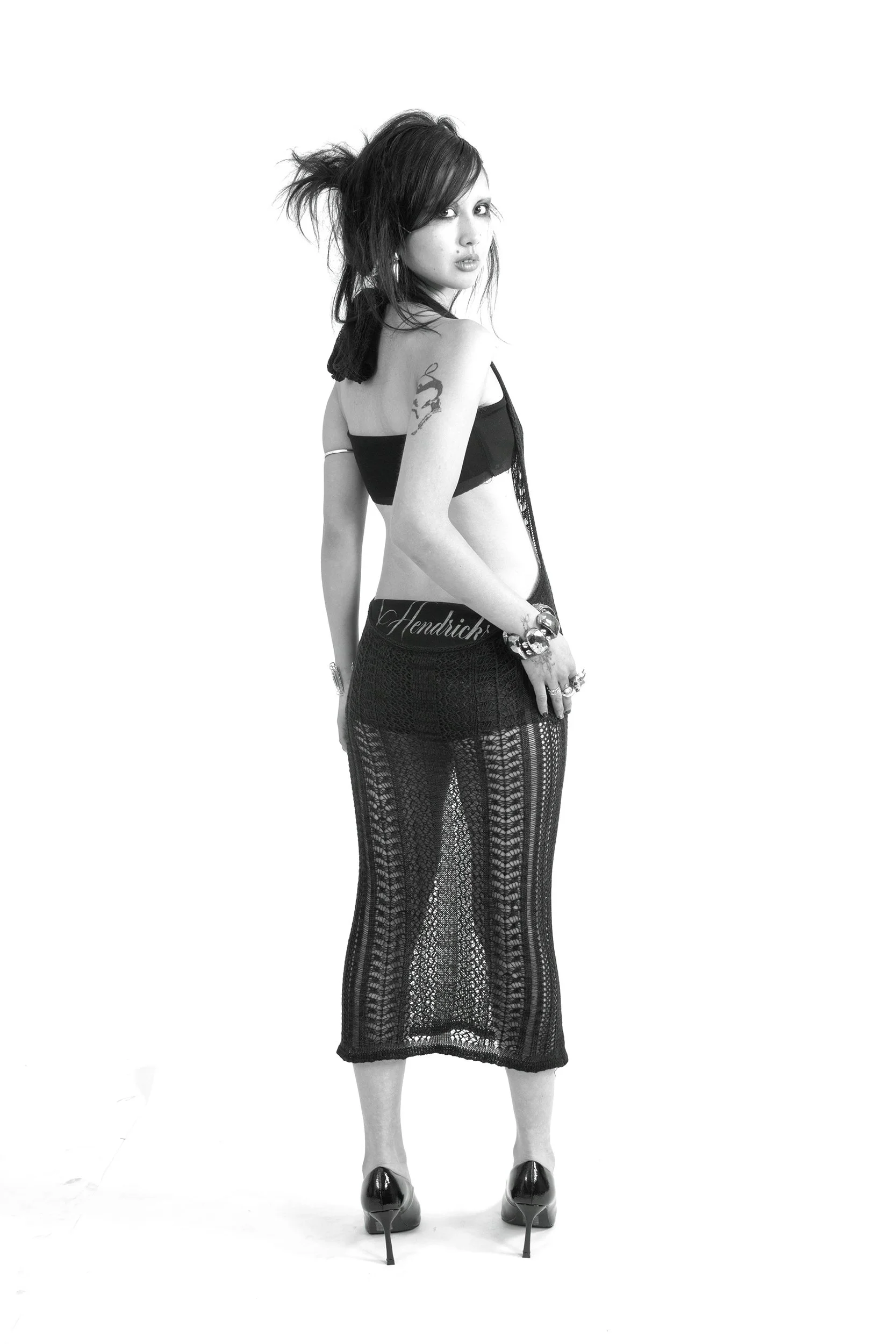 A woman in a black mesh dress, high heels, with a tattoo on her shoulder, standing with her back turned and looking over her shoulder."