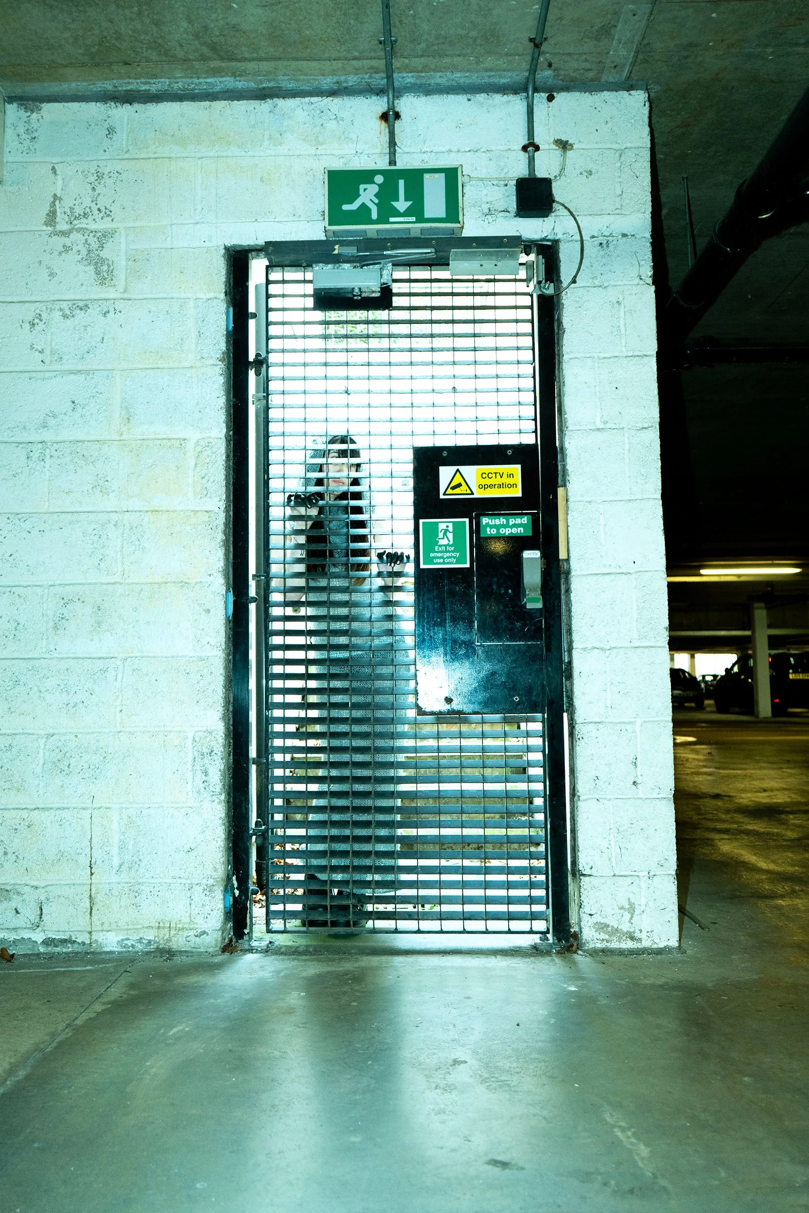 A secured gate with a green emergency exit sign above it, featuring a running figure and downward arrow, in an underground parking garage with a woman taking a photo in the reflection.
