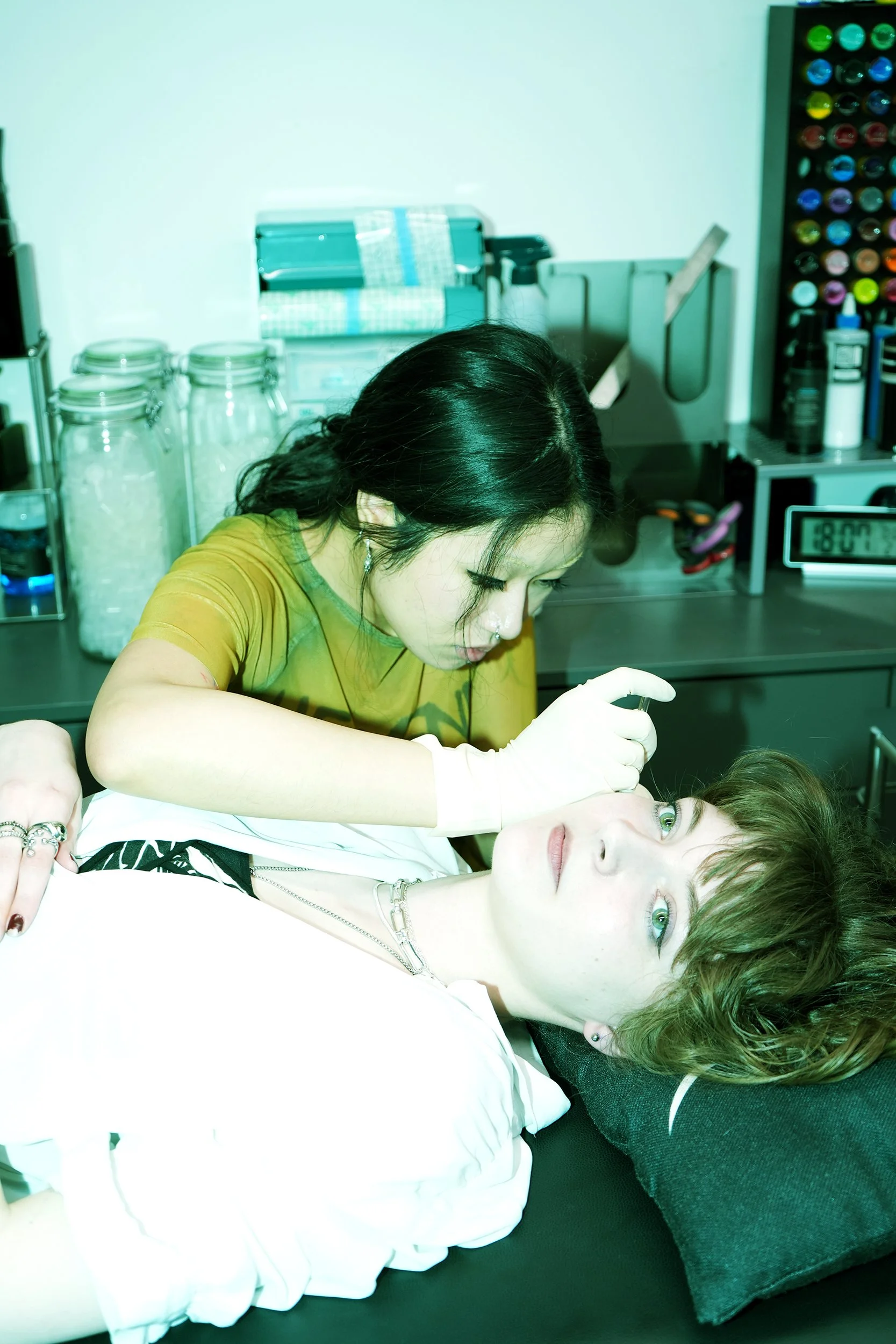 A person with short light brown hair and green eyes lying on a medical examination table, receiving a tattoo or similar procedure from a woman with black hair, wearing a yellow-green shirt and white gloves, in a medical or tattoo studio setting.