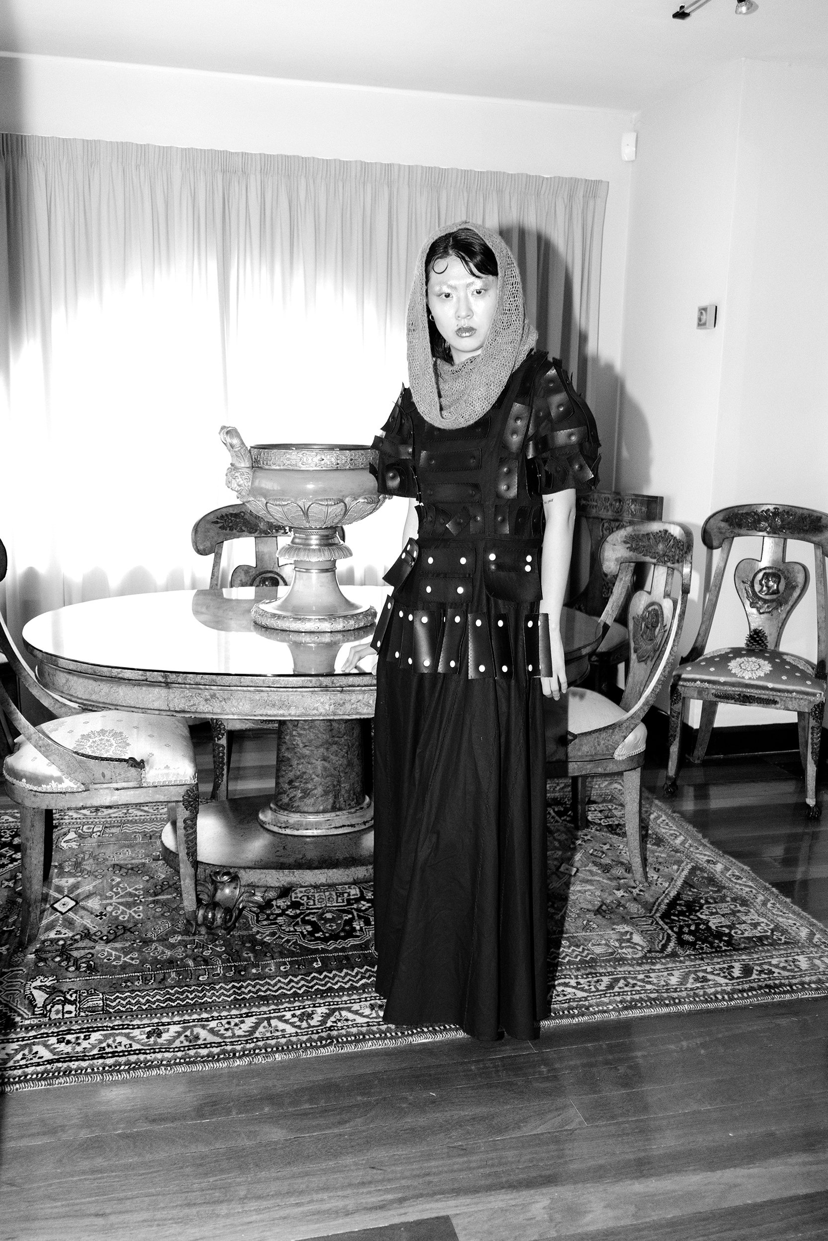 A woman with dark hair and a bonnet, wearing a dress with a leather-like top, stands in a dining room with a table, ornate chairs, a large decorative bowl, and an oriental rug.