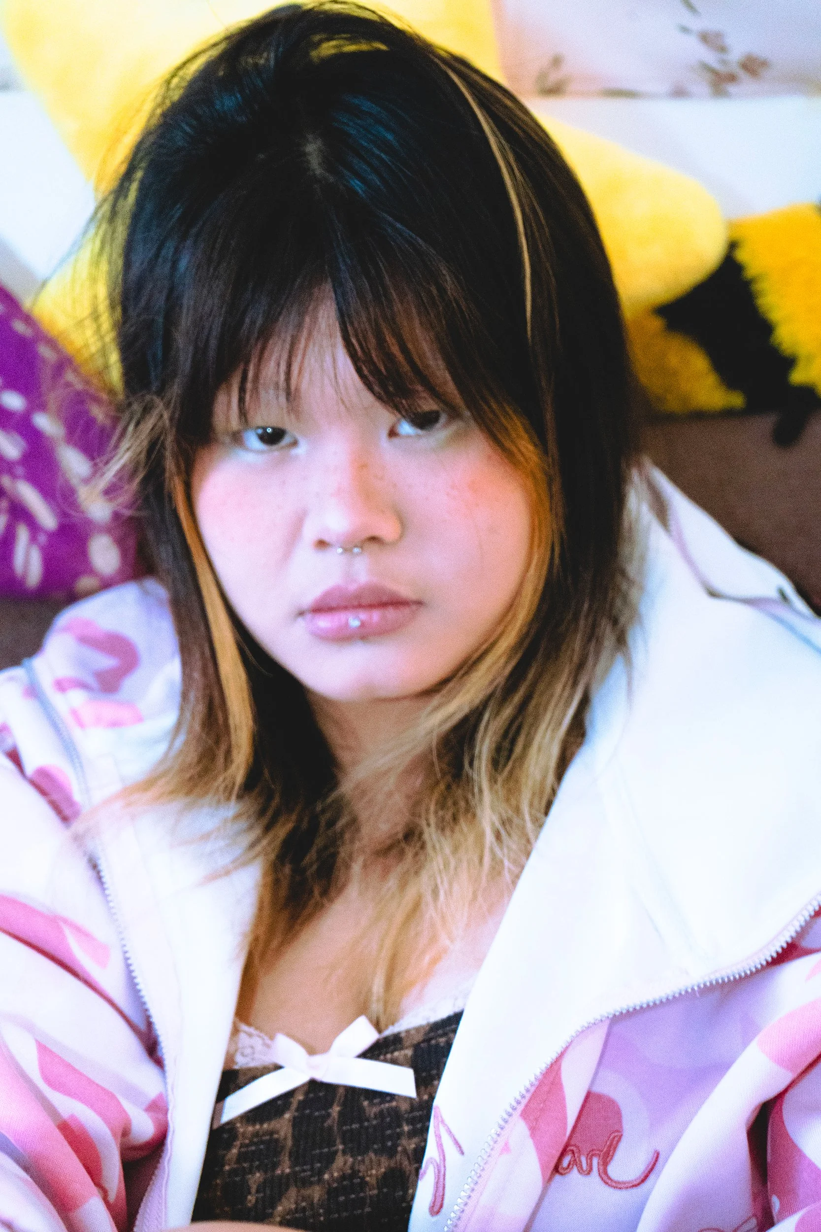 A woman with black and light brown hair lying on a bed, wearing a pink and white patterned jacket with a white bow and brown checkered top underneath, surrounded by yellow pillows.