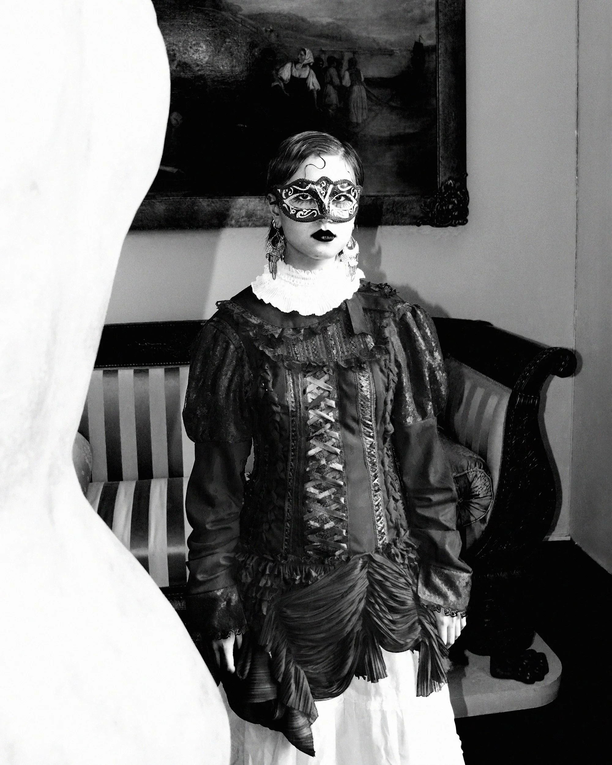 A woman with short hair wearing a decorative mask, ornate earrings, a ruffled collar, and a patterned dress, sitting on a draped fabric in front of a framed painting of people by a body of water.