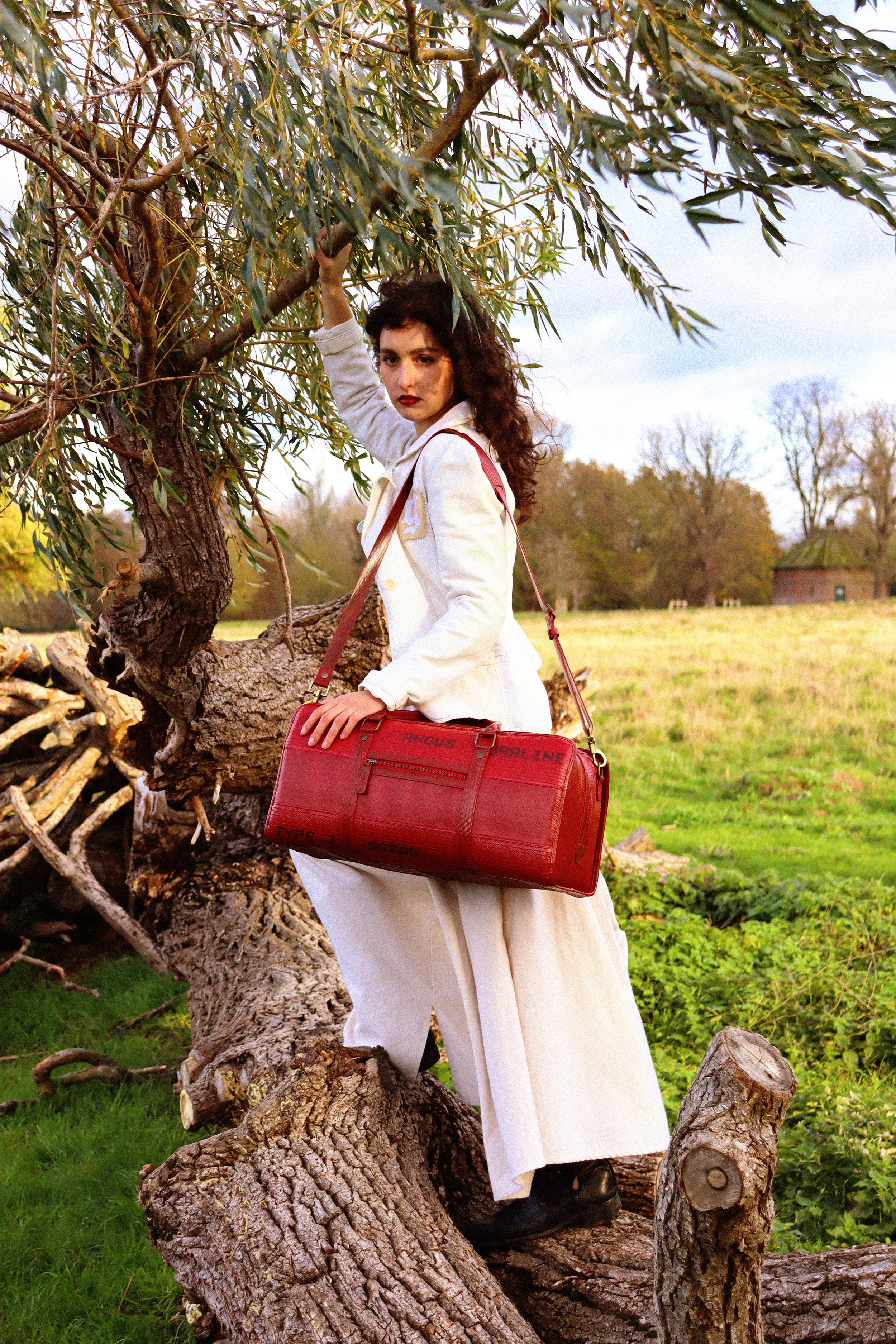 A woman with dark curly hair and red lipstick standing outdoors, leaning against a tree with twisted branches on a grassy field. She is wearing a white coat, white pants, black boots, and carrying a large red bag.
