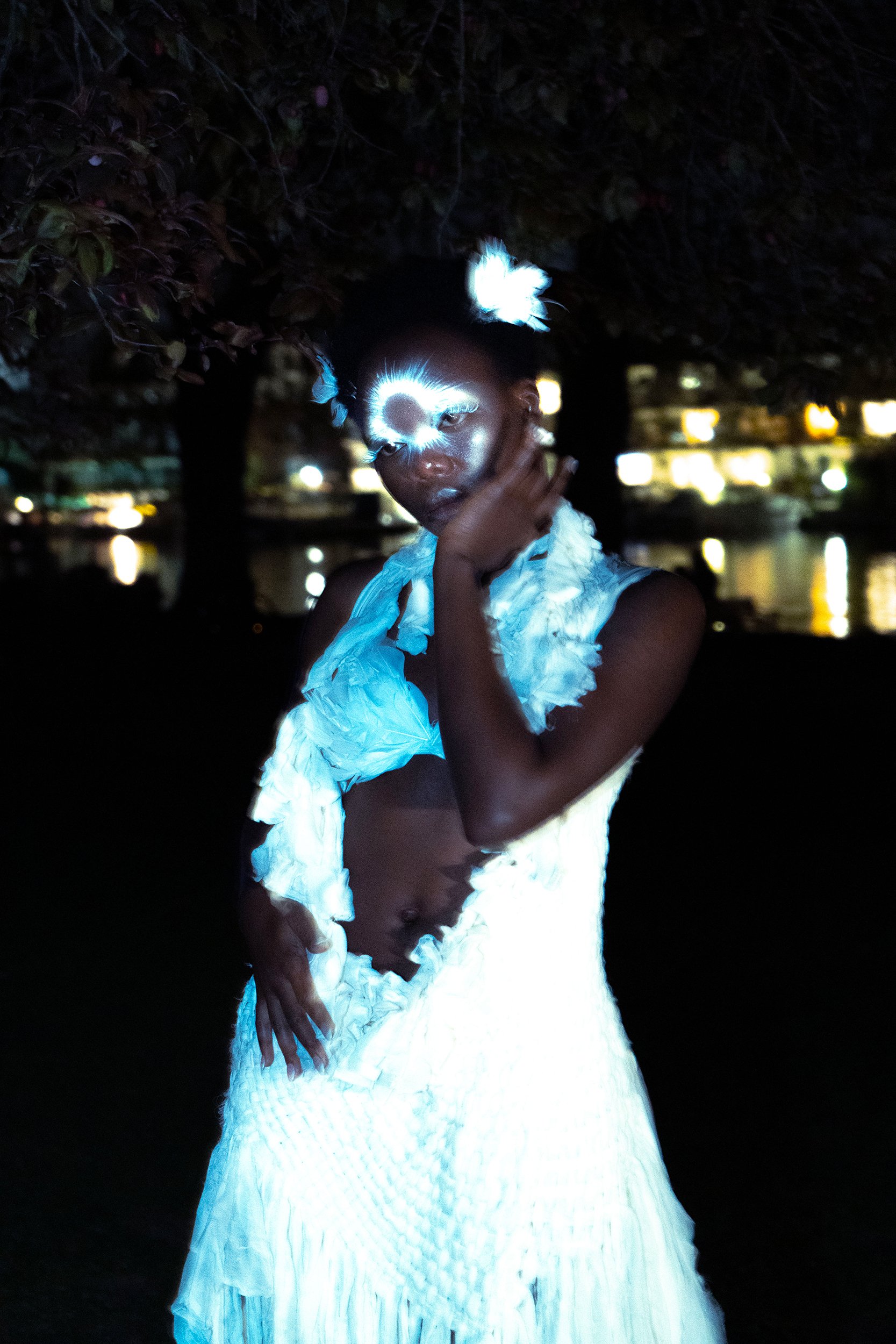 A person with glowing facial features and ears, wearing a white ruffled outfit, standing outdoors at night near water with blurred lights in the background.