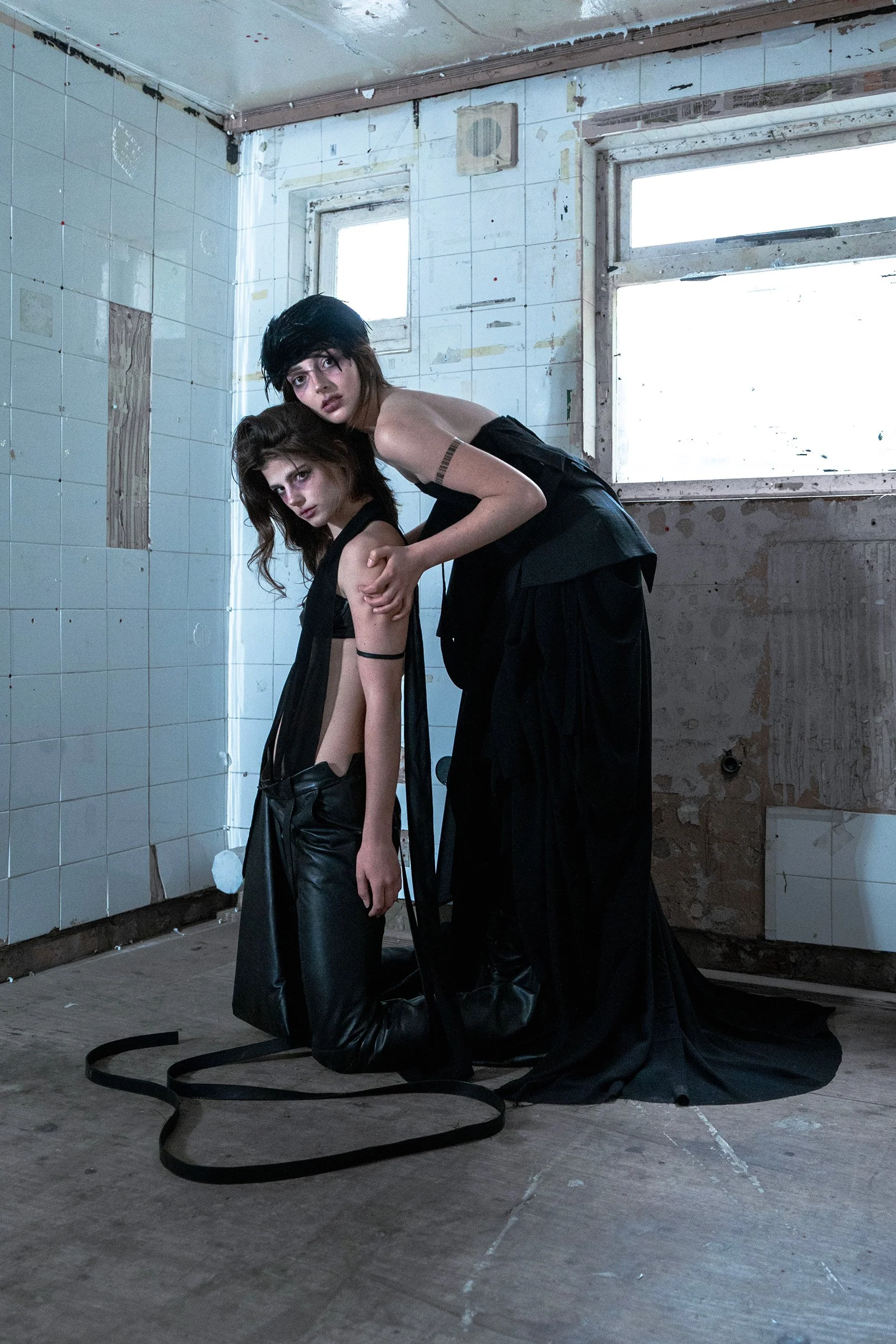 Two women with dark hair and makeup posing in a rundown, unfinished room with tiled walls and a concrete floor. They are dressed in black clothes, with one woman leaning on the other.