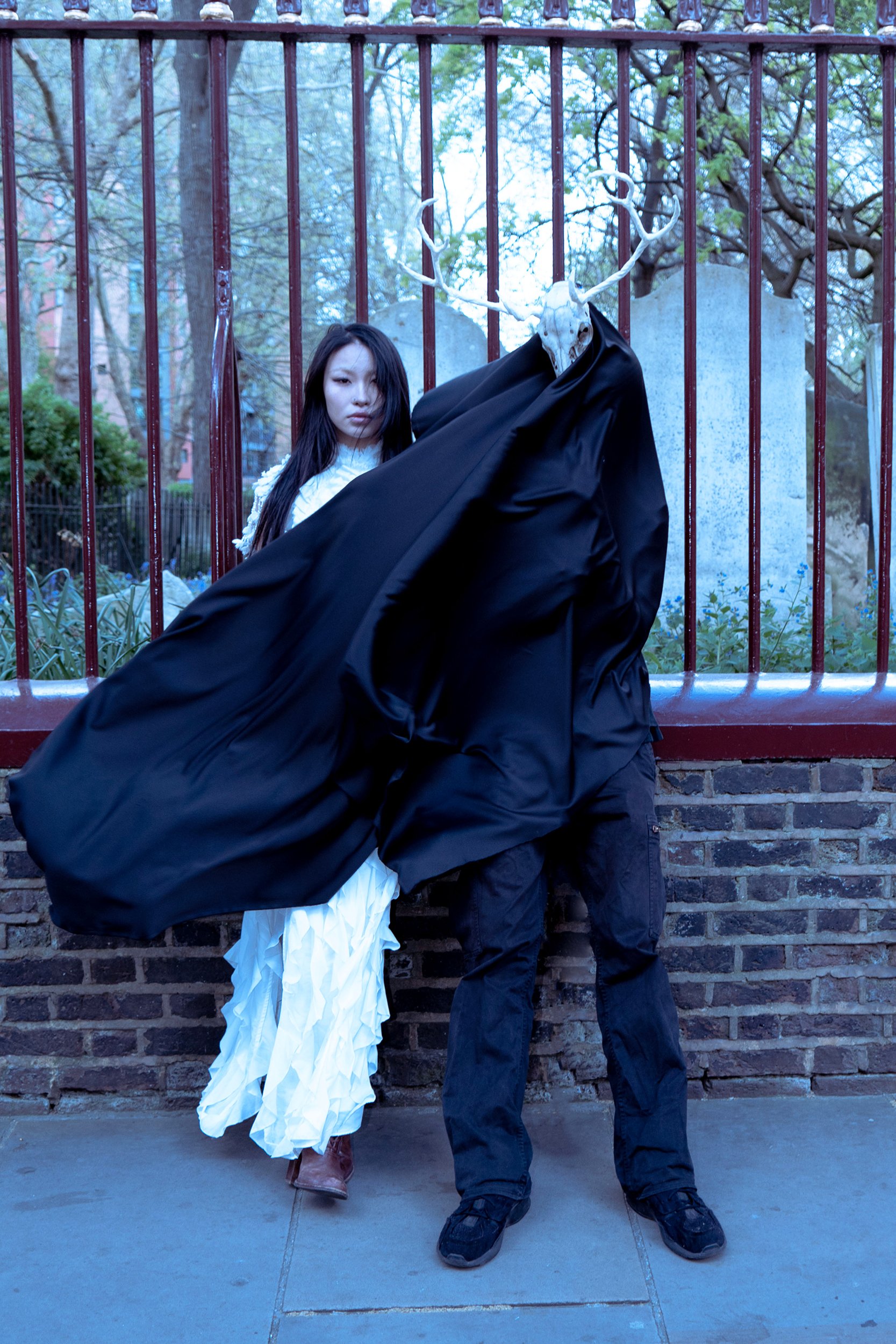 A woman in white standing next to a figure with a skull mask and antlers, dressed in black robes, standing behind a red metal fence in an outdoor area with trees in the background.