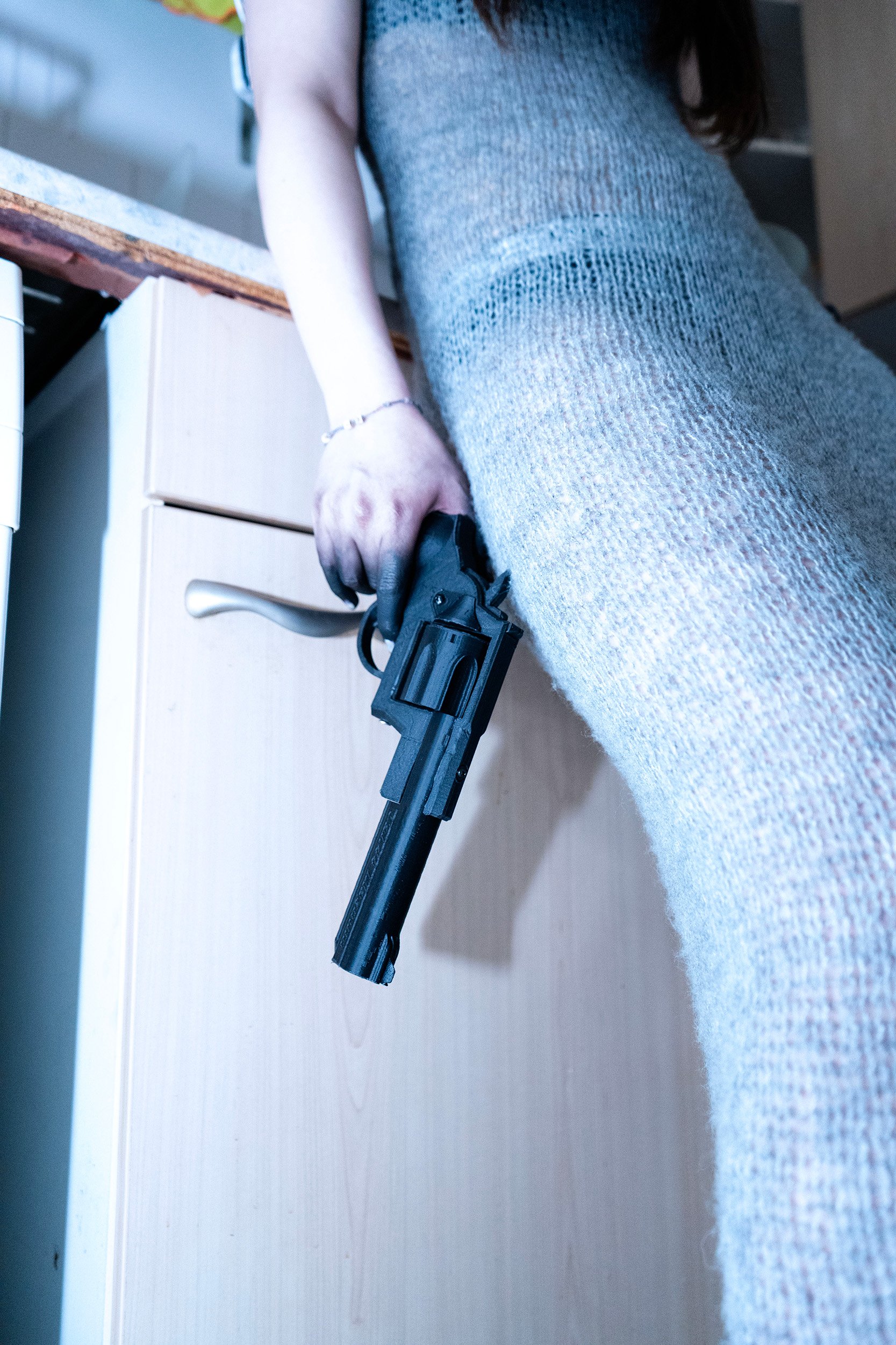 Person holding a black handgun with one hand, wearing a gray dress and a silver bracelet, standing in a room with a light-colored desk and a gray cabinet.