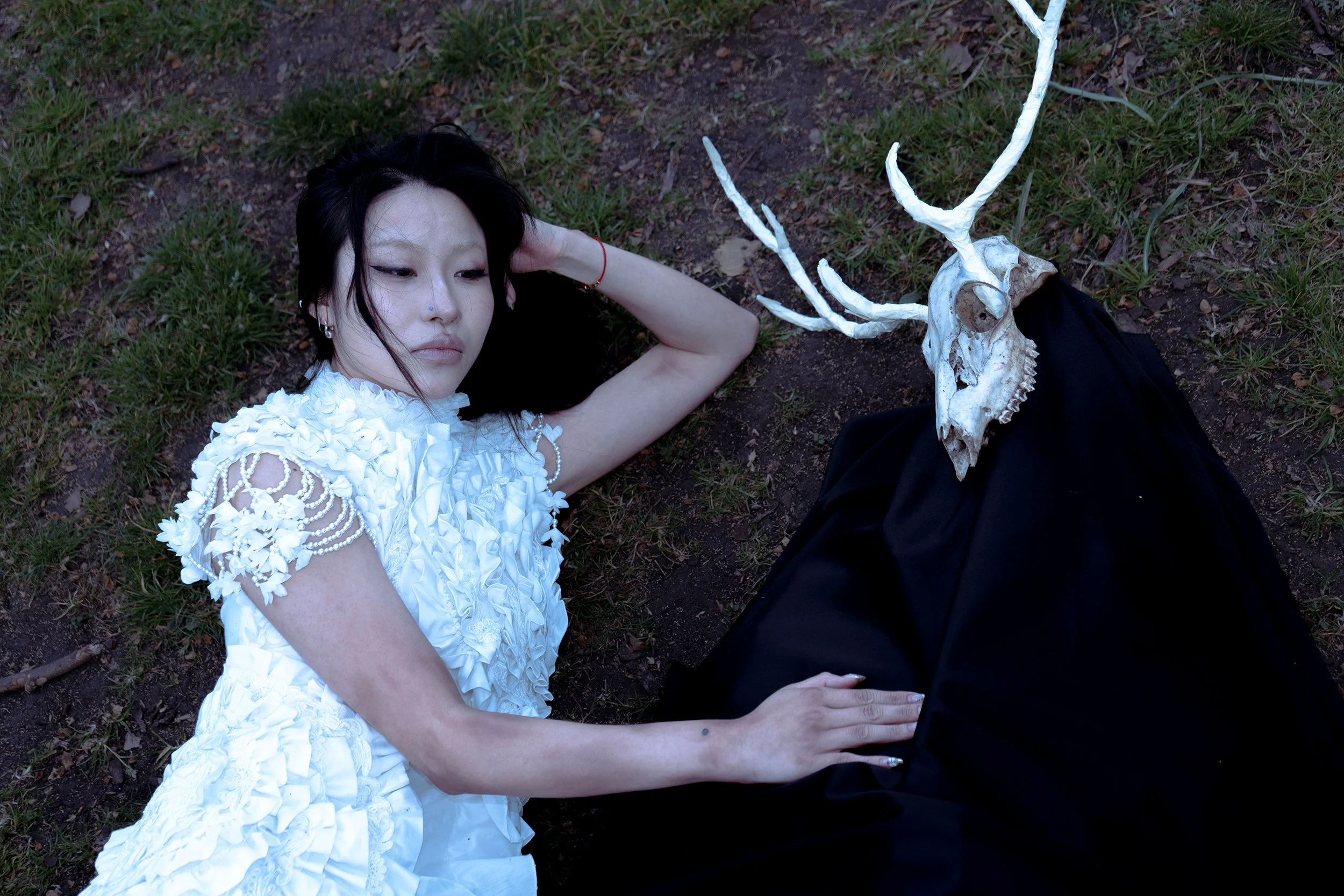 A woman wearing a white dress lying on the ground beside a deer skull with antlers.