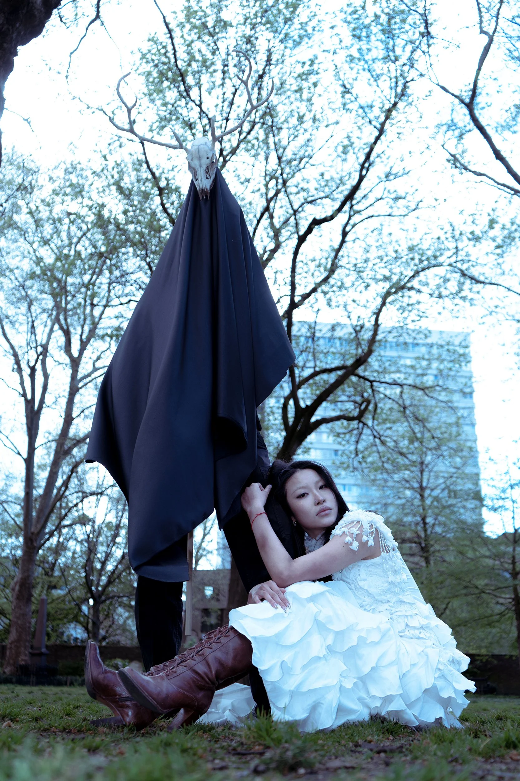 A young woman in a white, ruffled dress with lace details, brown knee-high lace-up boots, and red bracelets, is seated on the grass in a park. She has long dark hair and a calm expression, resting her head on her hand. Nearby, a standing figure is dressed in dark clothing with a large black cape, and wears a skull mask with antlers. The background features leafless trees and a tall modern building, suggesting an urban park setting.