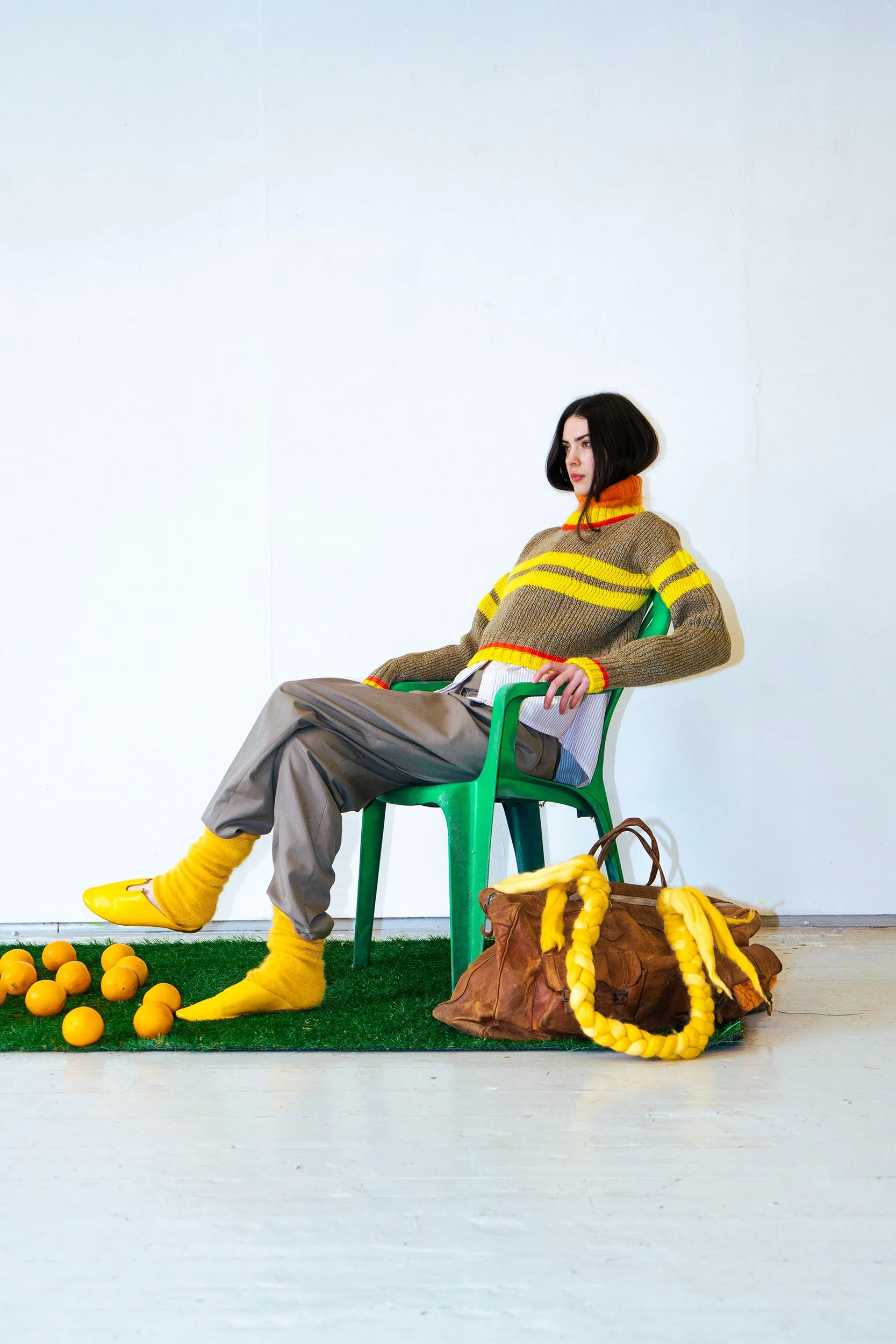 Woman with shoulder-length black hair sitting on a green plastic chair, wearing a brown and yellow striped sweater, gray baggy pants, yellow shoes and yellow socks, with a yellow scarf around her neck, against a plain white wall. She has a serious expression. There are yellow balls on green artificial grass and yellow shoes and a brown bag with yellow knitted loops on the floor.