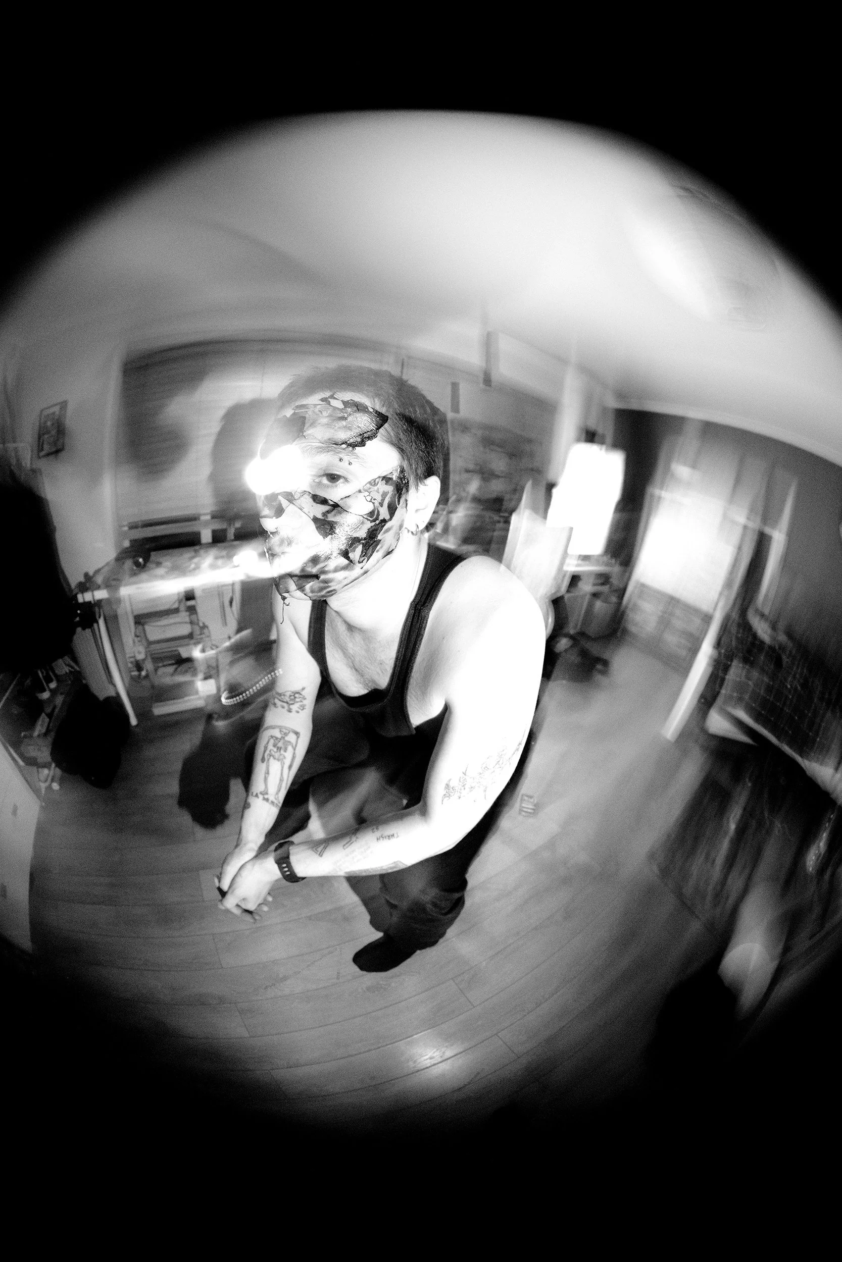 A person with tattoos on their arms and wearing a floral face mask and black tank top taking a selfie in a fish-eye lens effect mirror in a room with wooden floors and furniture.