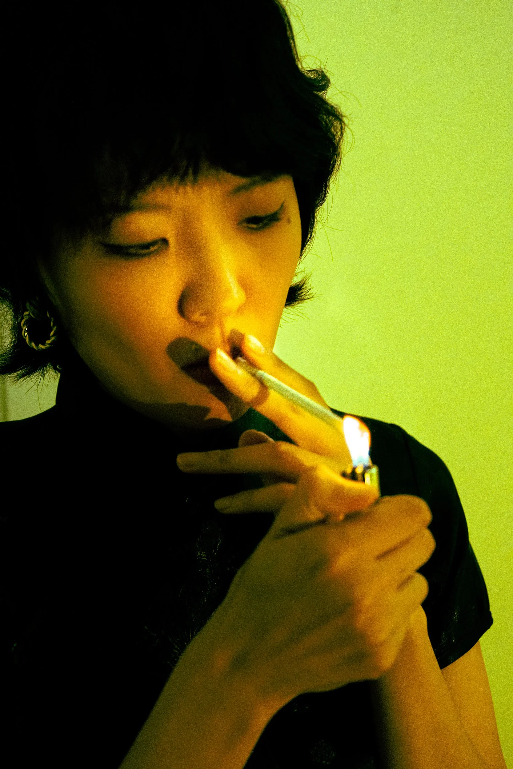A person with short black hair and earrings is lighting a cigarette with a lighter against a green background.