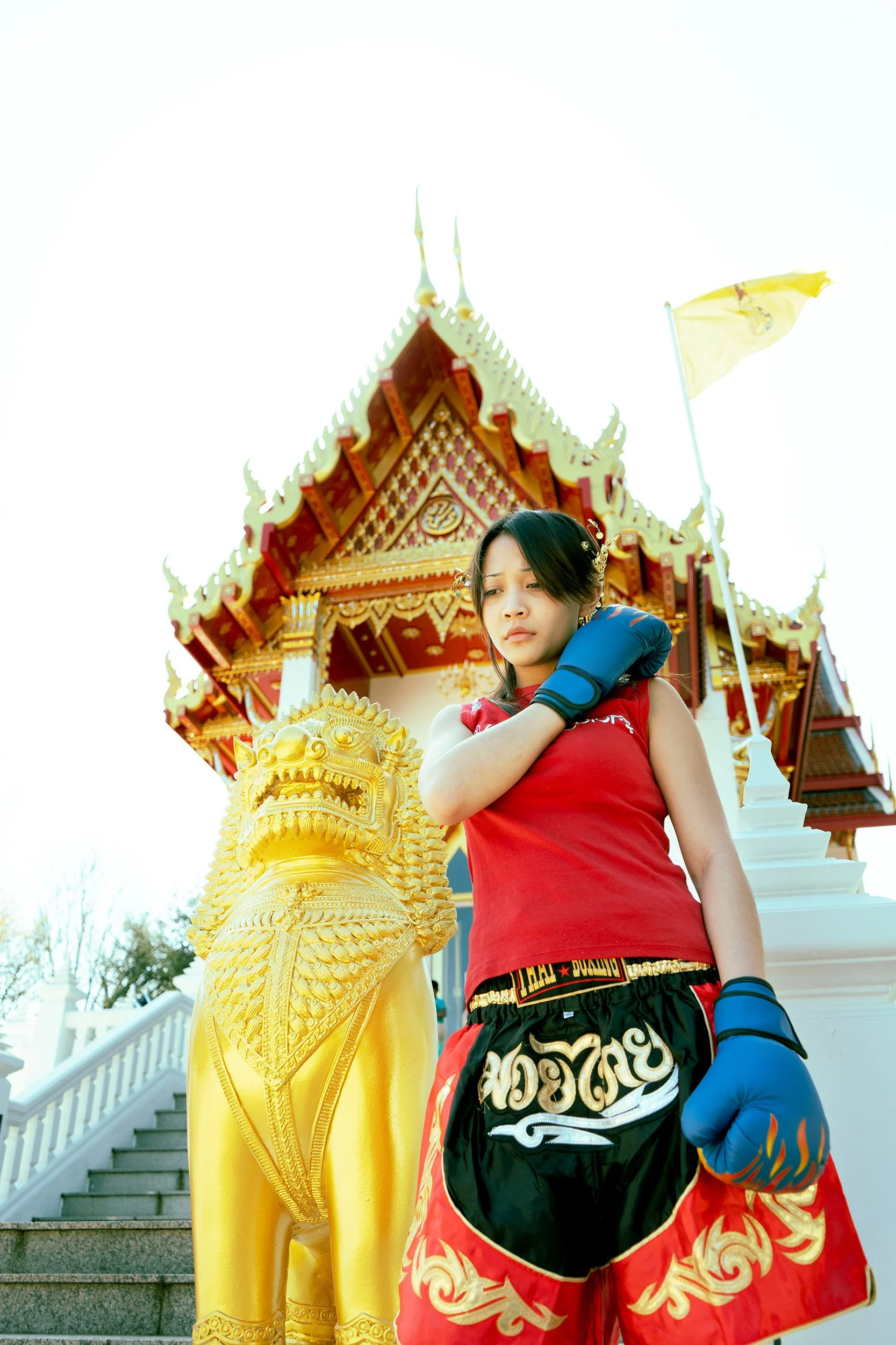 A woman dressed in a red and black boxing outfit with blue gloves and golden ornate trim, standing in front of a traditional Thai temple with intricate decorations and a white flag, on a staircase.
