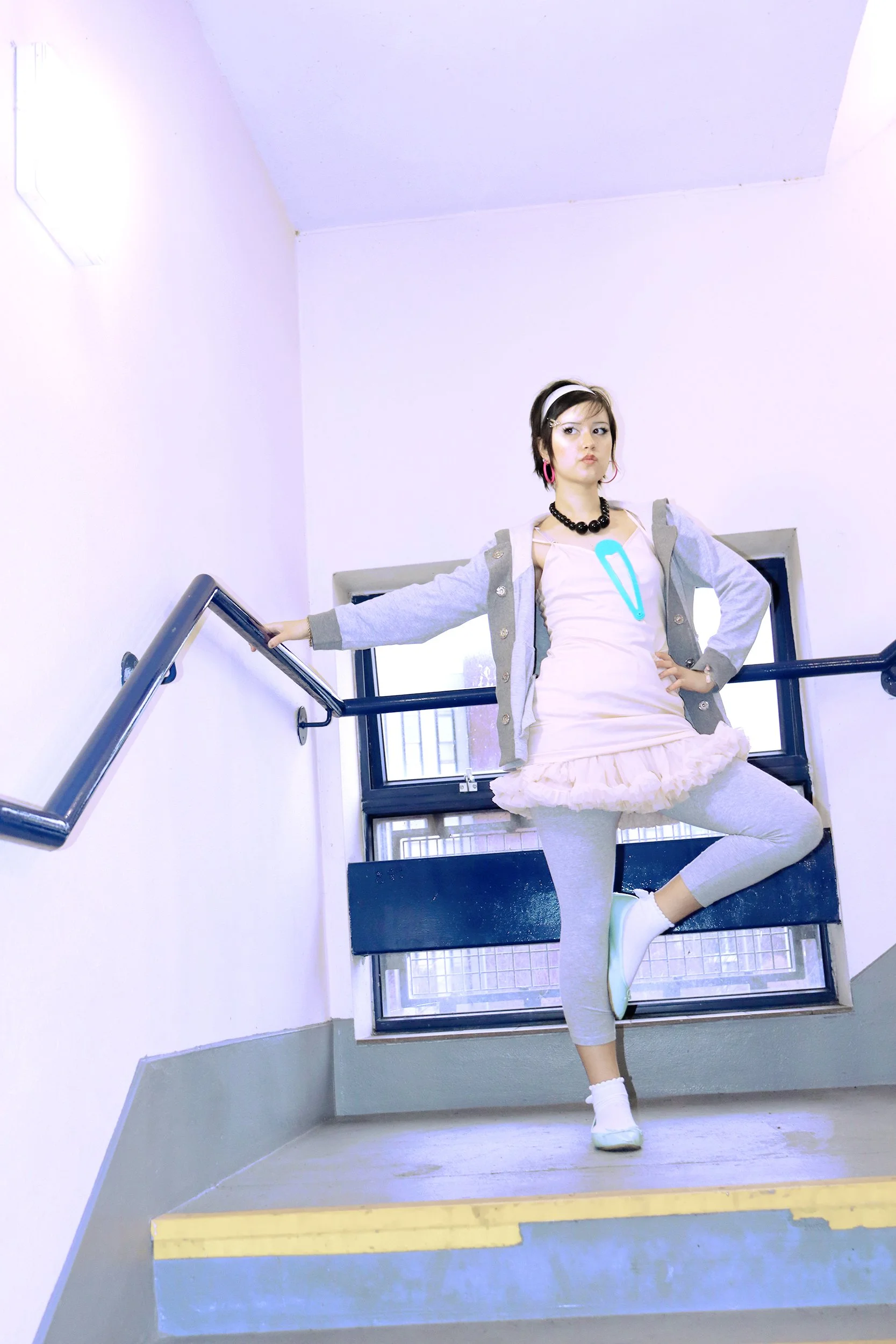 A young woman standing on a staircase in a building, striking a pose with one arm extended and one hand on her hip. She is wearing a gray hoodie, a pink dress with ruffled hem, black beads necklace, pink earrings, white shoes, and a headband. The staircase has yellow and gray steps and metal railings, with windows in the background allowing natural light to fill the space.