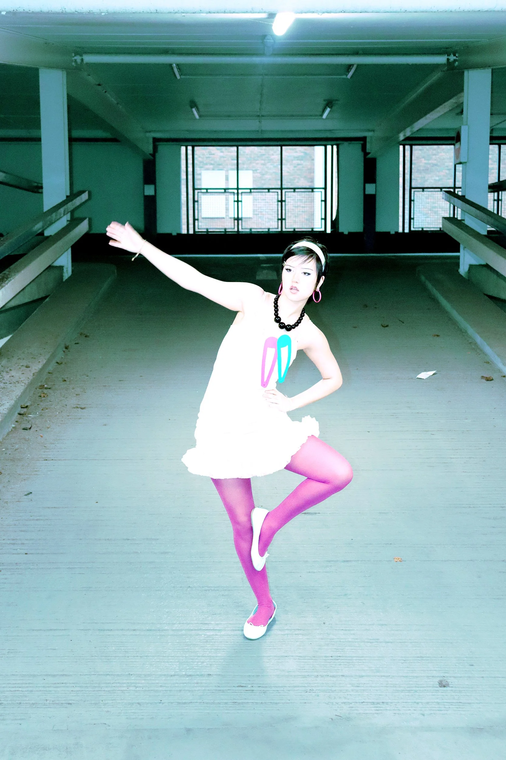 A woman in a white dress with colorful earrings and a headband is posing in an outdoor parking garage, with her left arm extended outward and right hand on her hip, standing on one leg with the other bent and foot pointed.