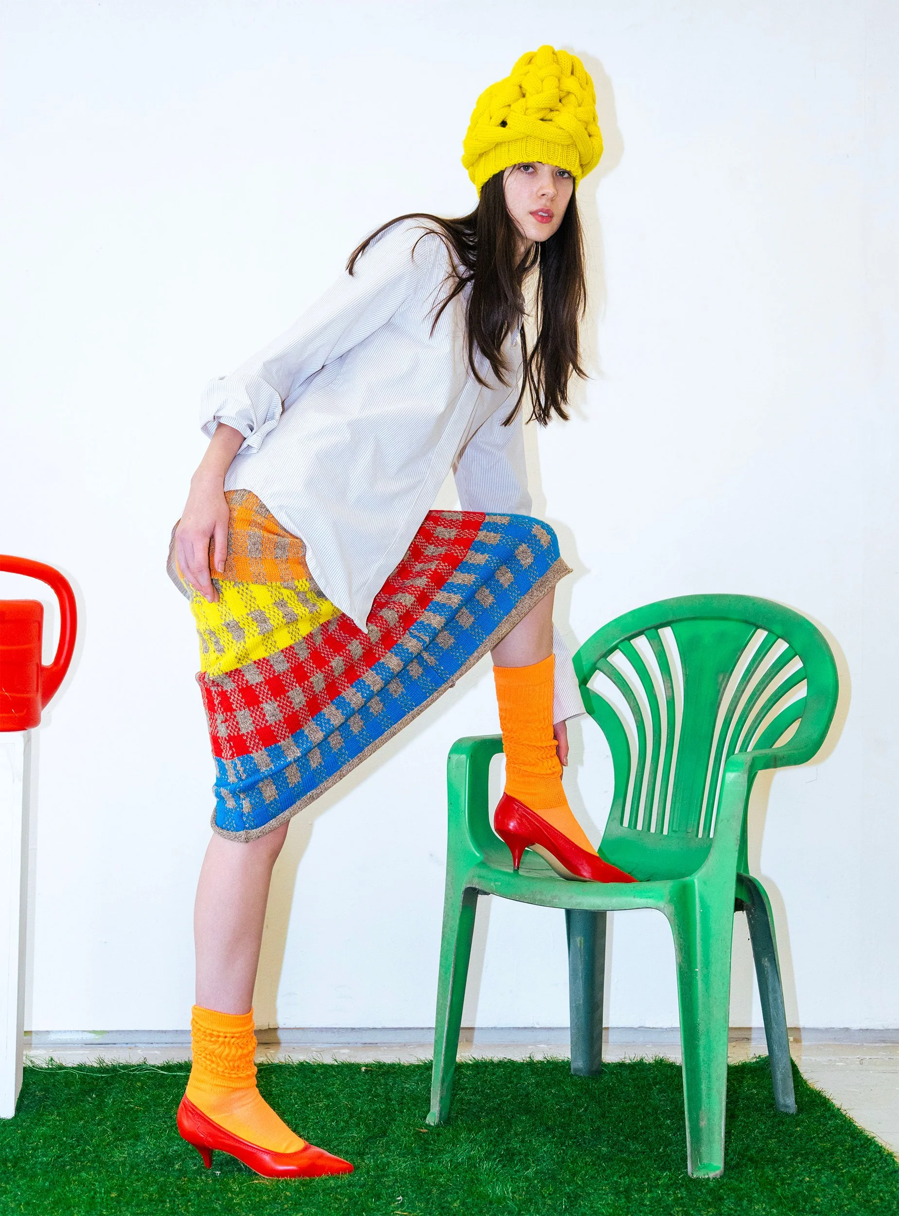 Woman wearing an oversized yellow knit hat, white shirt, colorful plaid skirt, orange socks, red high heels, standing next to a green plastic chair inside a room with polka dot carpet.