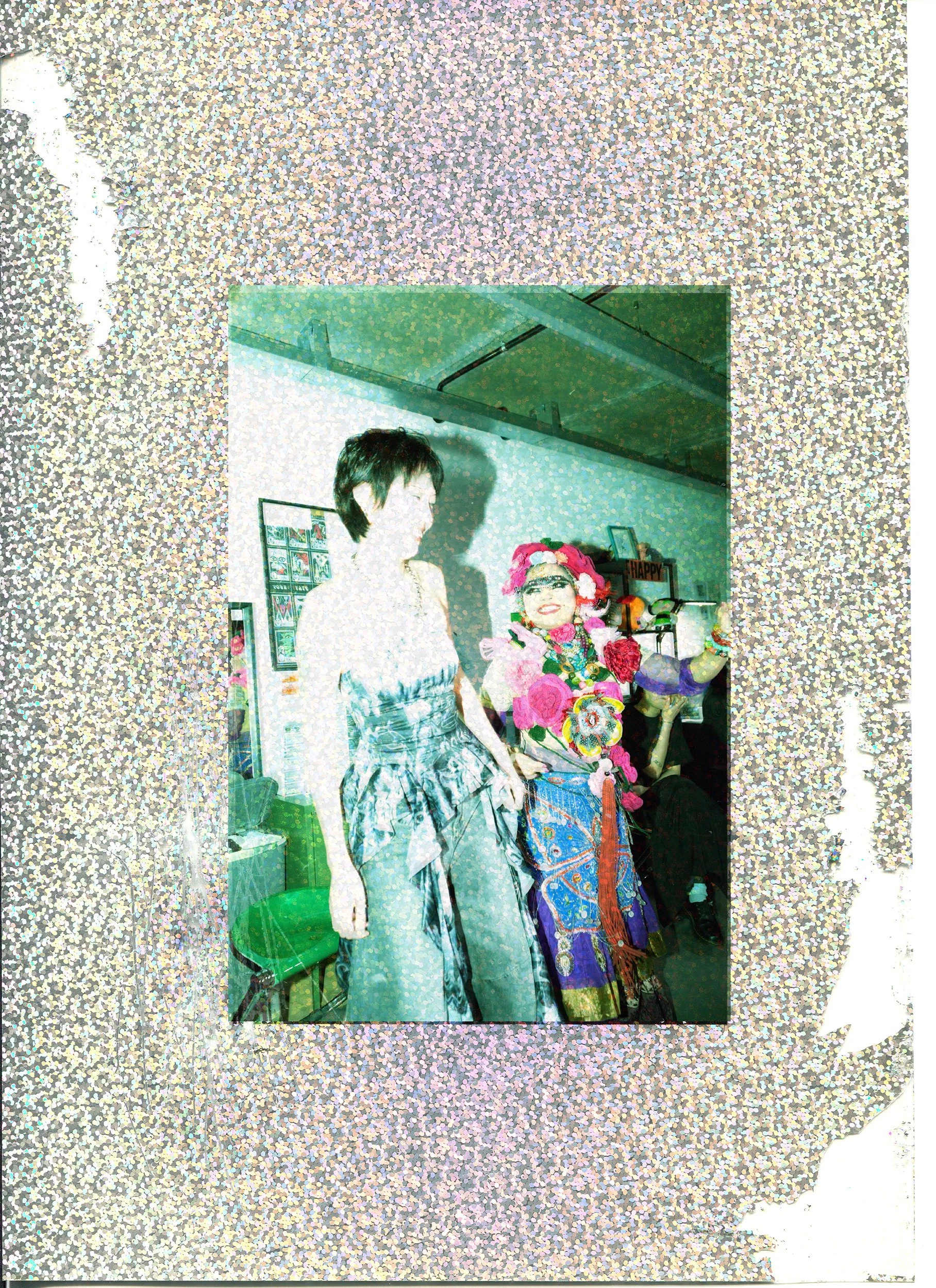 Photo of two women dressed in colorful, traditional, cultural attire, standing indoors, with a background decorated with framed pictures and a small sign reading 'HAPPY'.