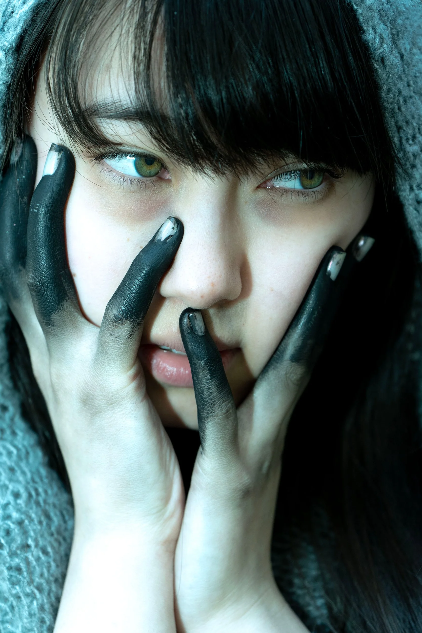 Close-up of a person's face with green eyes, black hair, and light skin, holding their face with both hands painted black, partially covering their face and eyes.