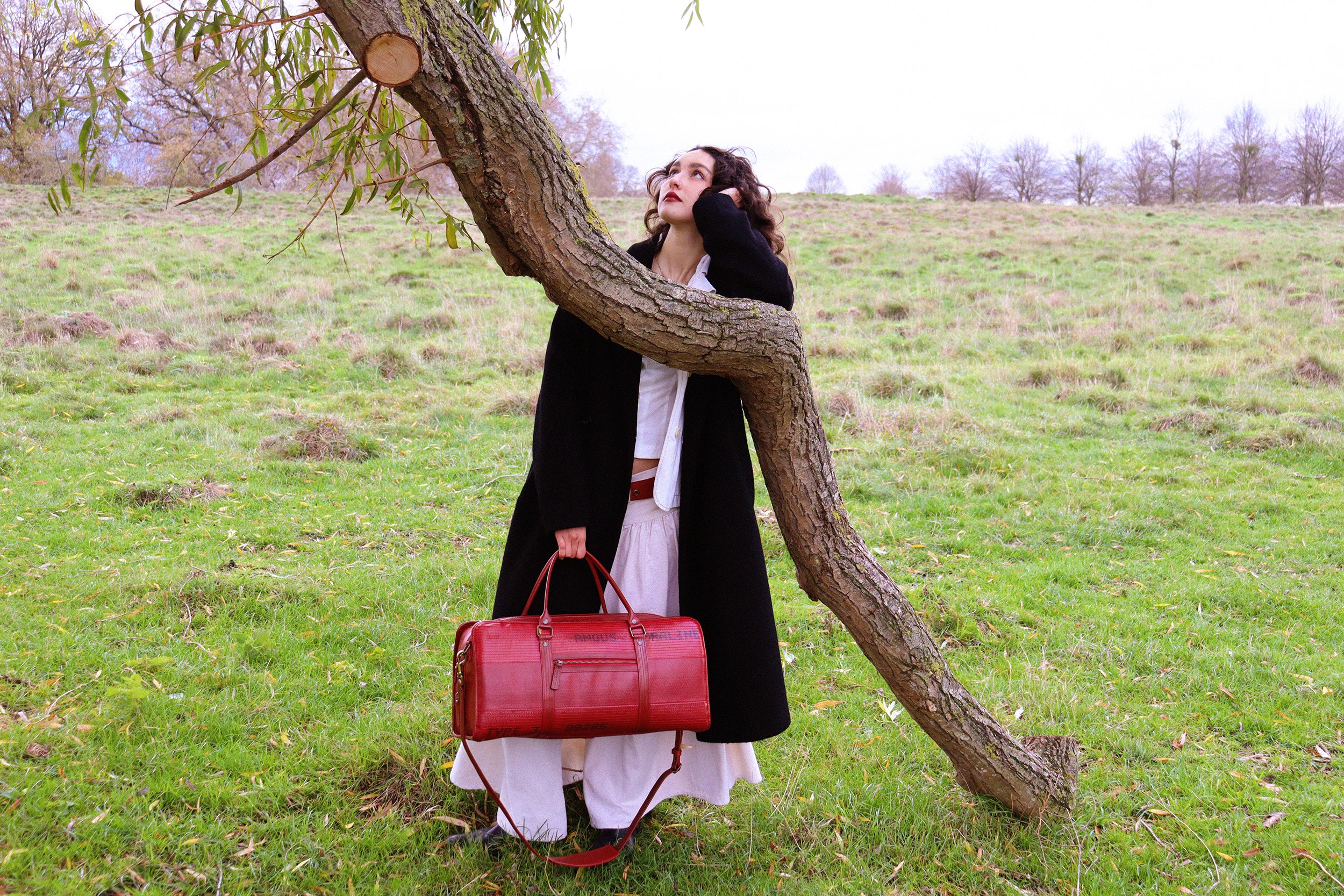 A woman with dark curly hair, dressed in a black coat and white outfit, stands in a grassy field with trees in the background. She rests her head on her hand, looking up, and holds a red handbag.