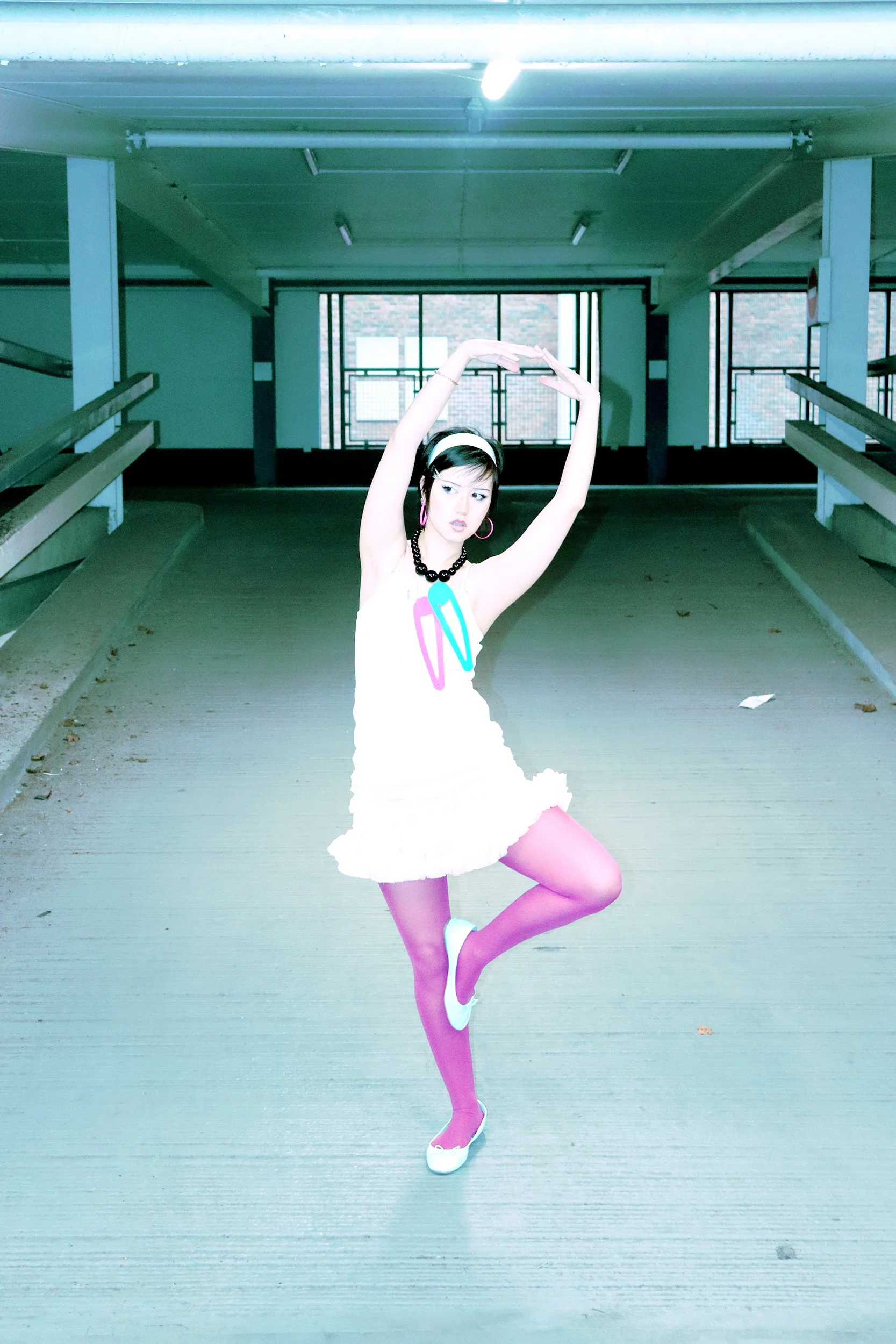 A woman with short dark hair, wearing a white headband, pink hoop earrings, a black beaded necklace, a white dress with colorful shapes, purple tights, and white shoes, poses in a parking garage with her arms raised above her head.