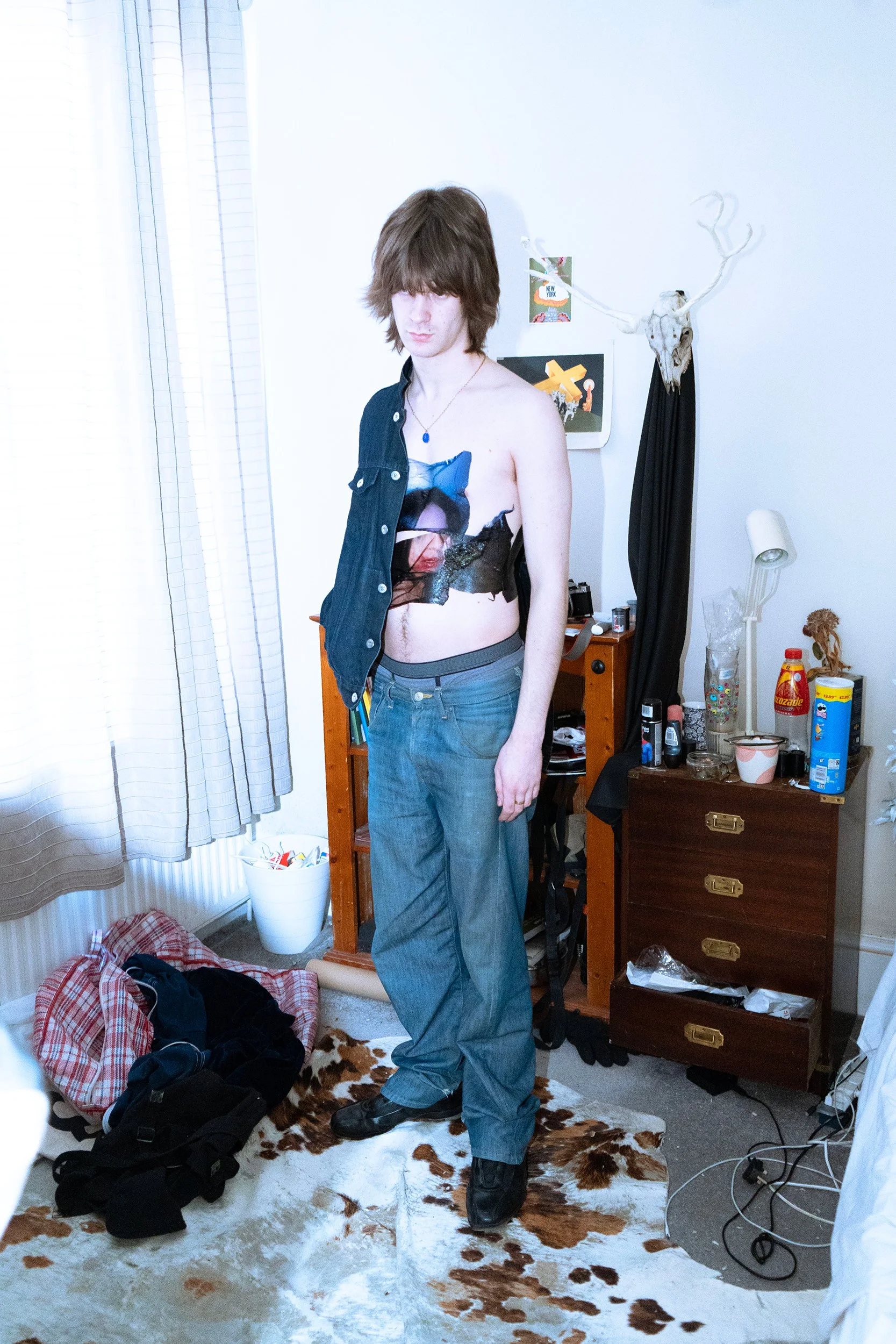 A young man with long brown hair is standing in a cluttered room, partially undressed and wearing baggy jeans and black boots, with a cowhide rug on the floor. The room has a white wall with a skull of a moose with antlers and some art posters behind him. There are various items on a small wooden dresser, including snacks, bottles, and accessories, and a laundry basket on the carpet, with clothes and other objects scattered around.