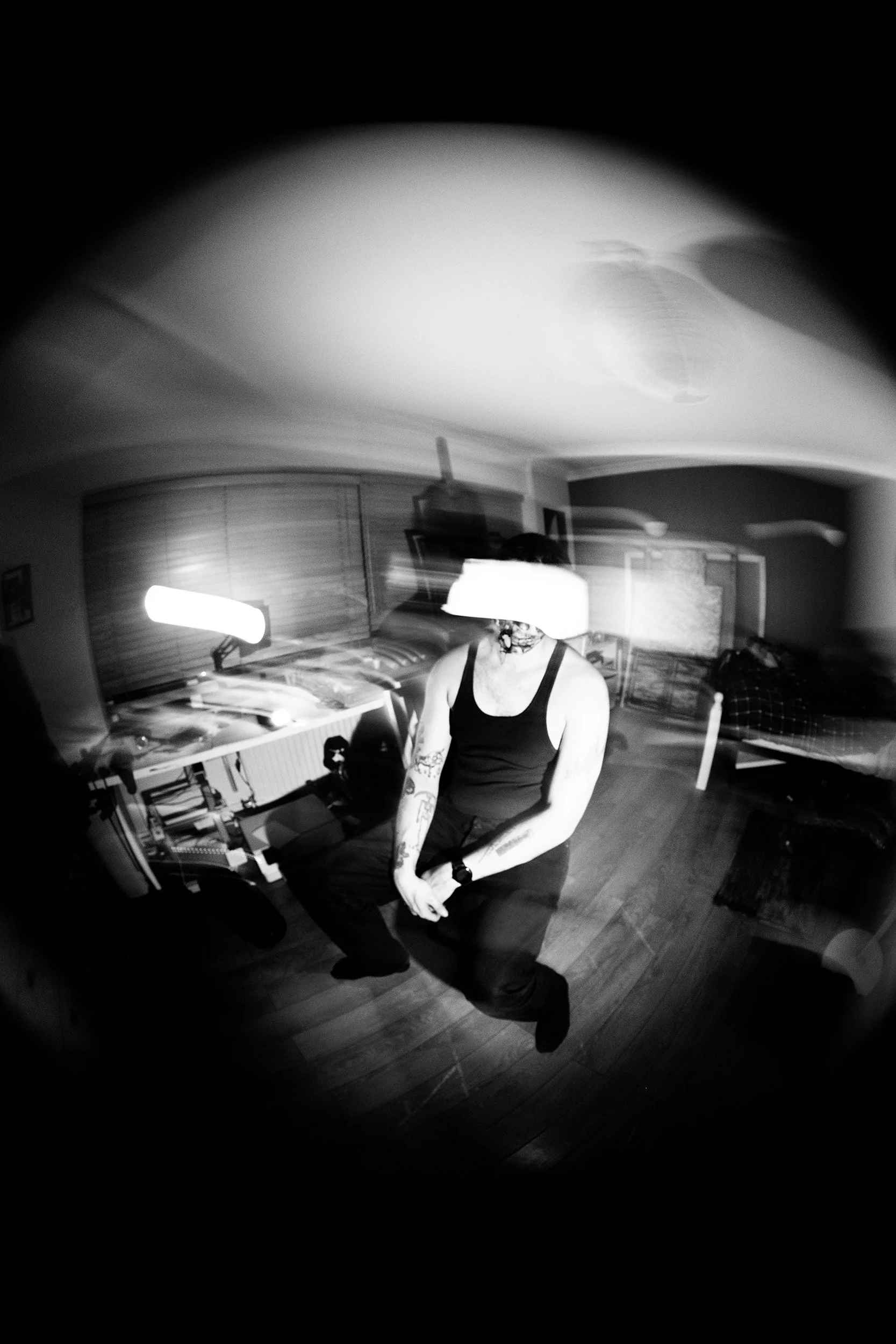 A person with tattoos on their right arm and wearing a tank top and glasses, sitting on the floor in a room with wooden flooring and a bookcase in the background.