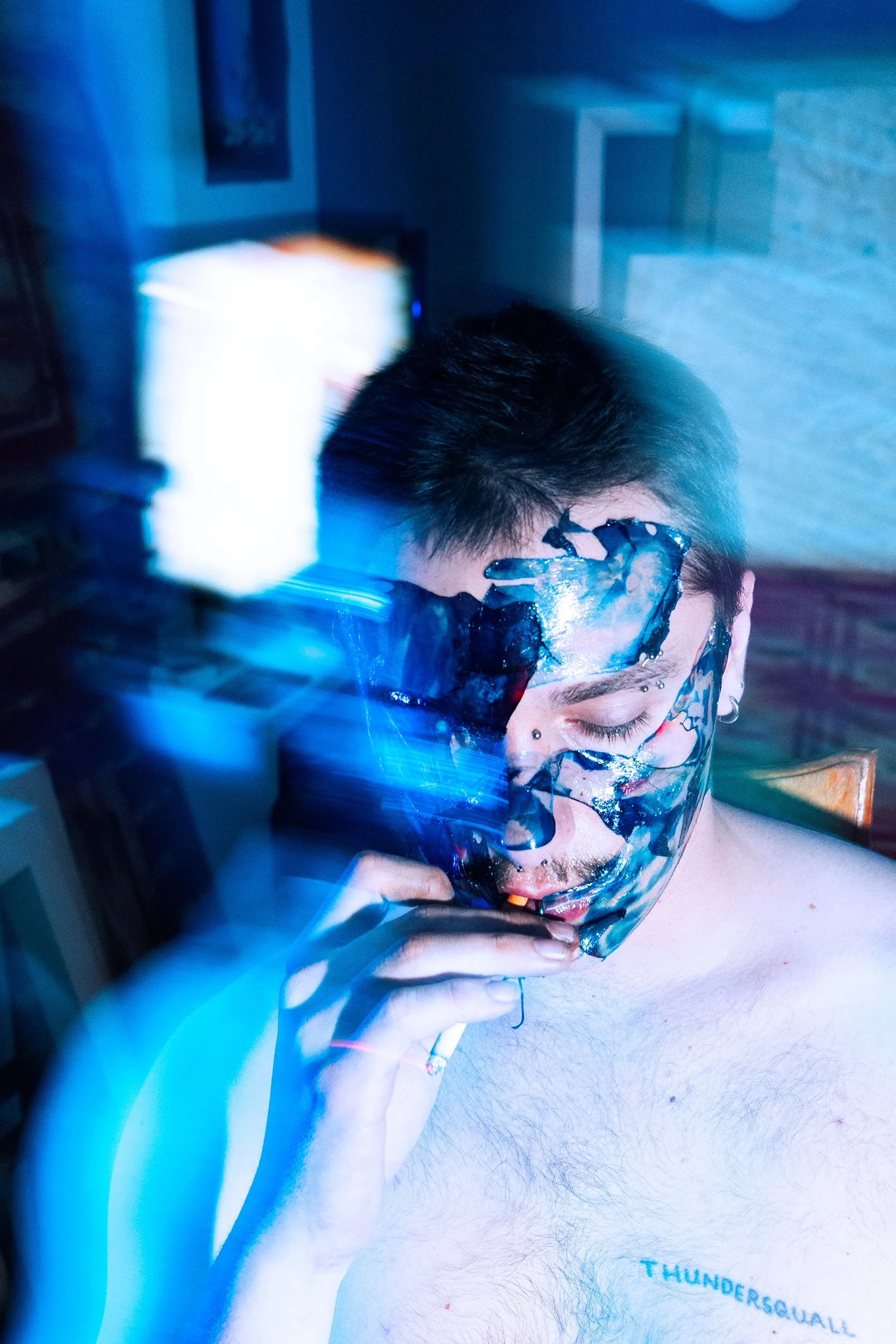 A young man with tattoos on his face and hand, with one eye closed and his head resting on his arm, which is placed on his chest. His shirt is partially visible with the word 'THUNDER' printed on it, and the background appears to be an indoor setting.