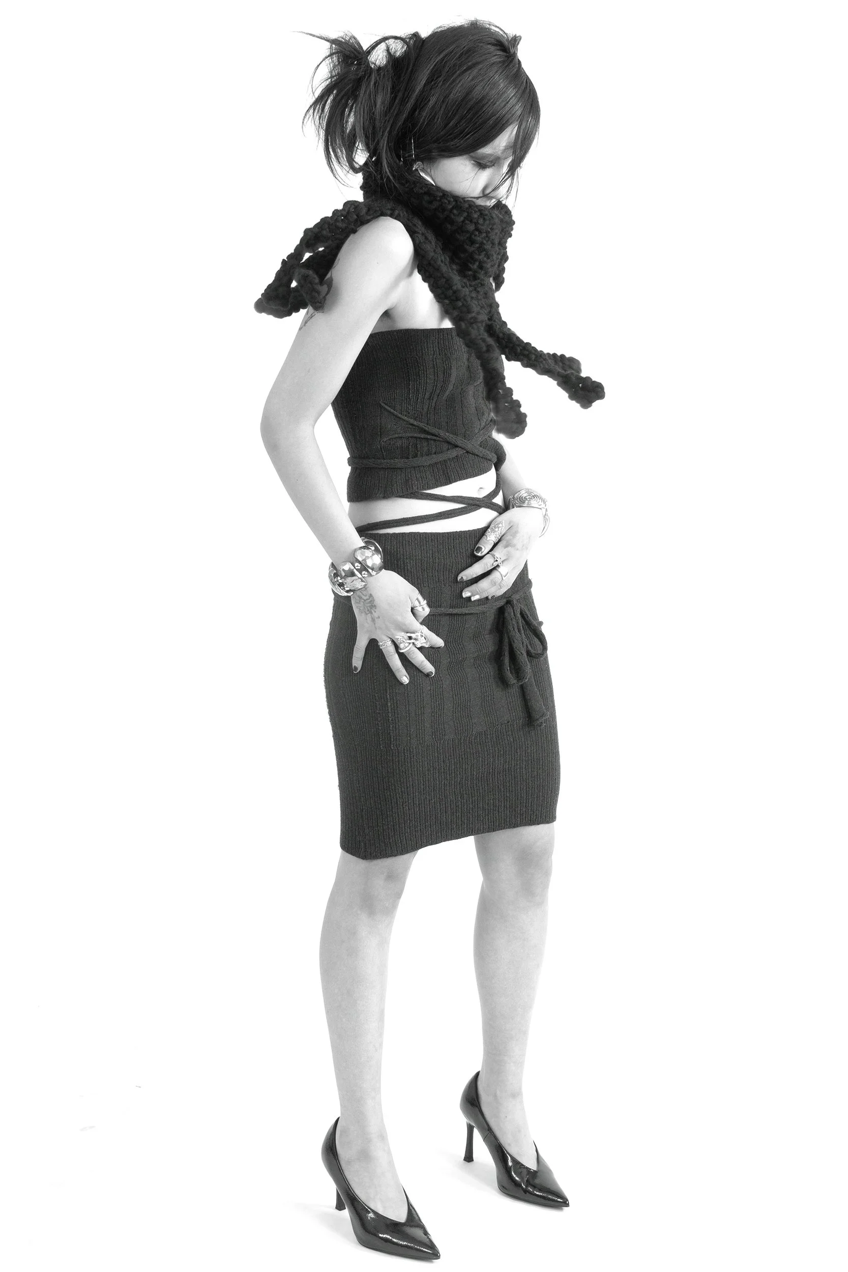 Black and white photo of a woman wearing high heels, a short skirt, and a sleeveless top, standing sideways with her head down.