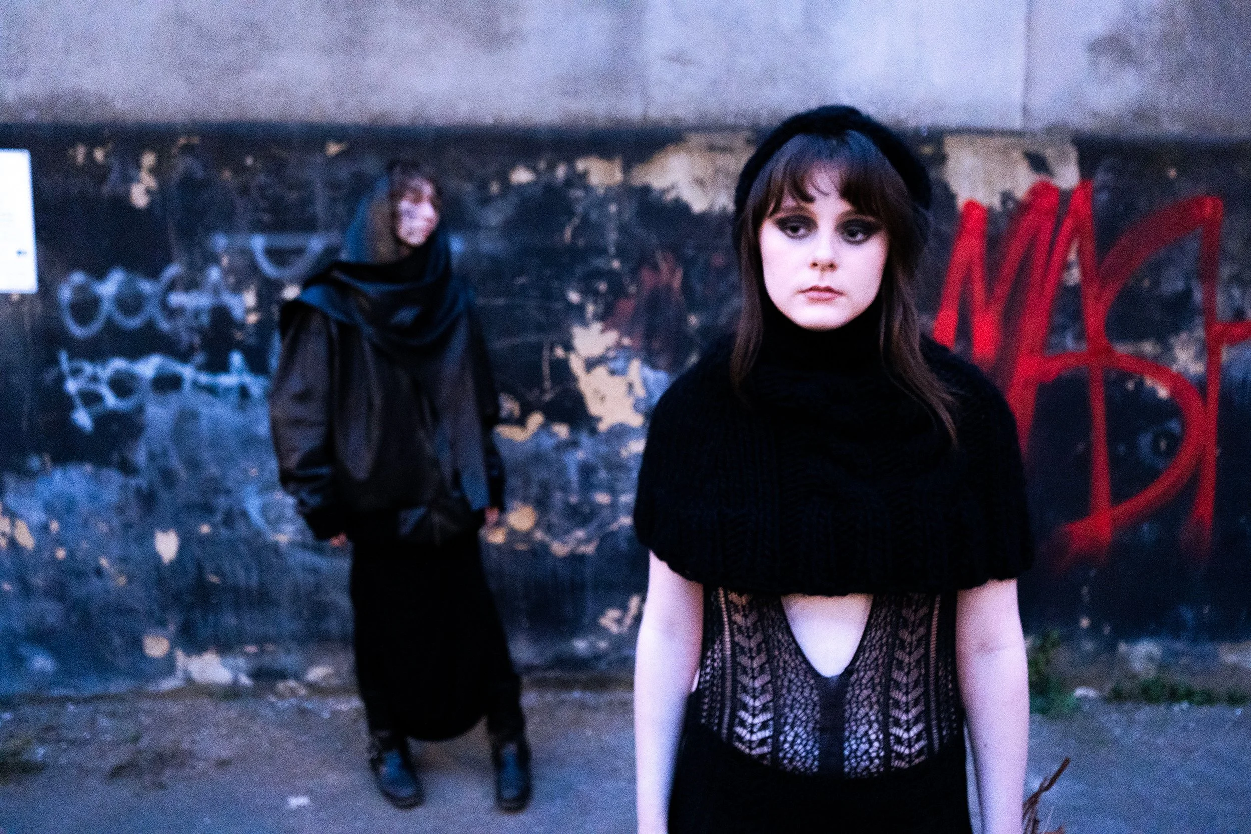 Two women standing in front of a graffiti-covered wall. The woman in the foreground has long brown hair, wearing a black hat and black lace dress with a deep neckline, looking pensively to the side. The second woman is in the background, wearing a dark jacket and a black headscarf, slightly out of focus.