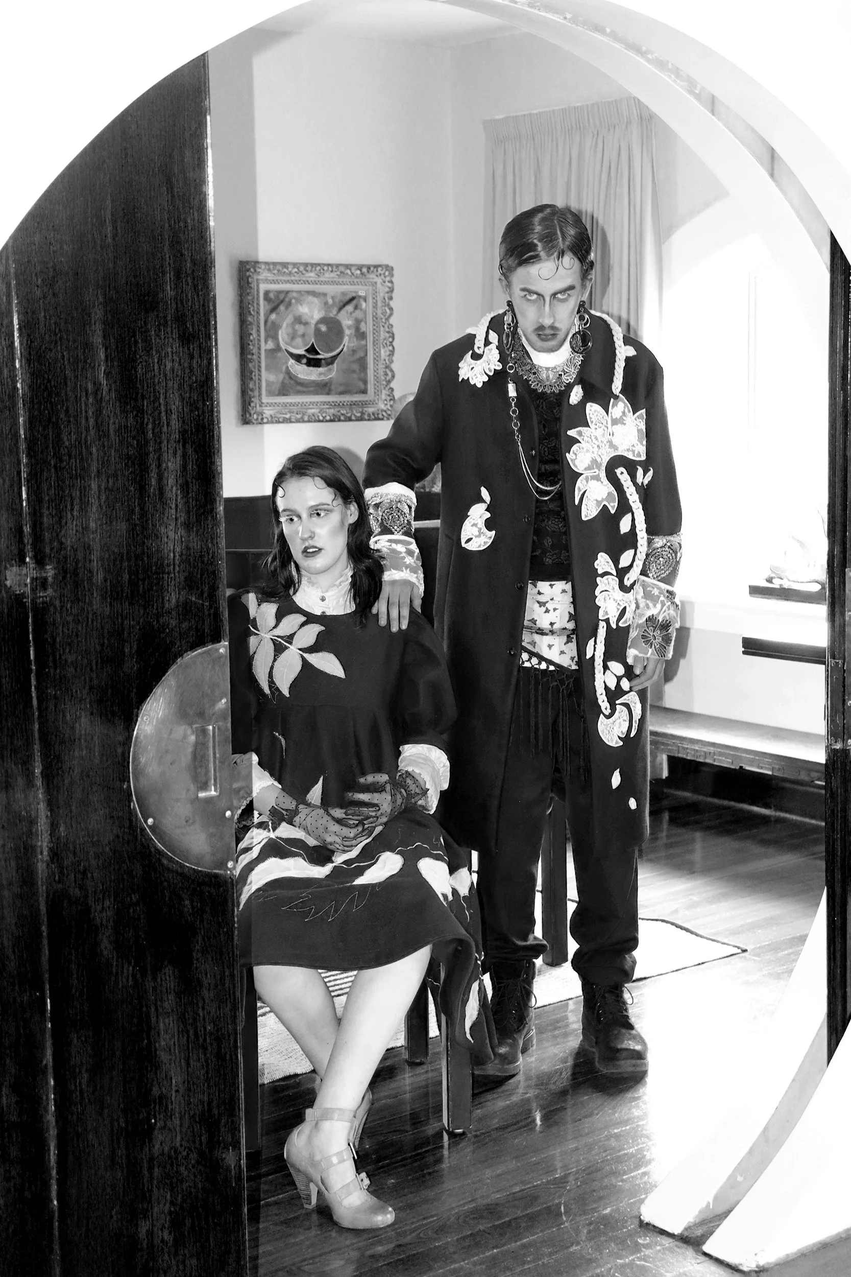 A black and white photo of a man standing next to a seated woman, both dressed in vintage, elaborate clothing with bold makeup, in a room with artwork and curtains, reflecting a stylized, dramatic scene.