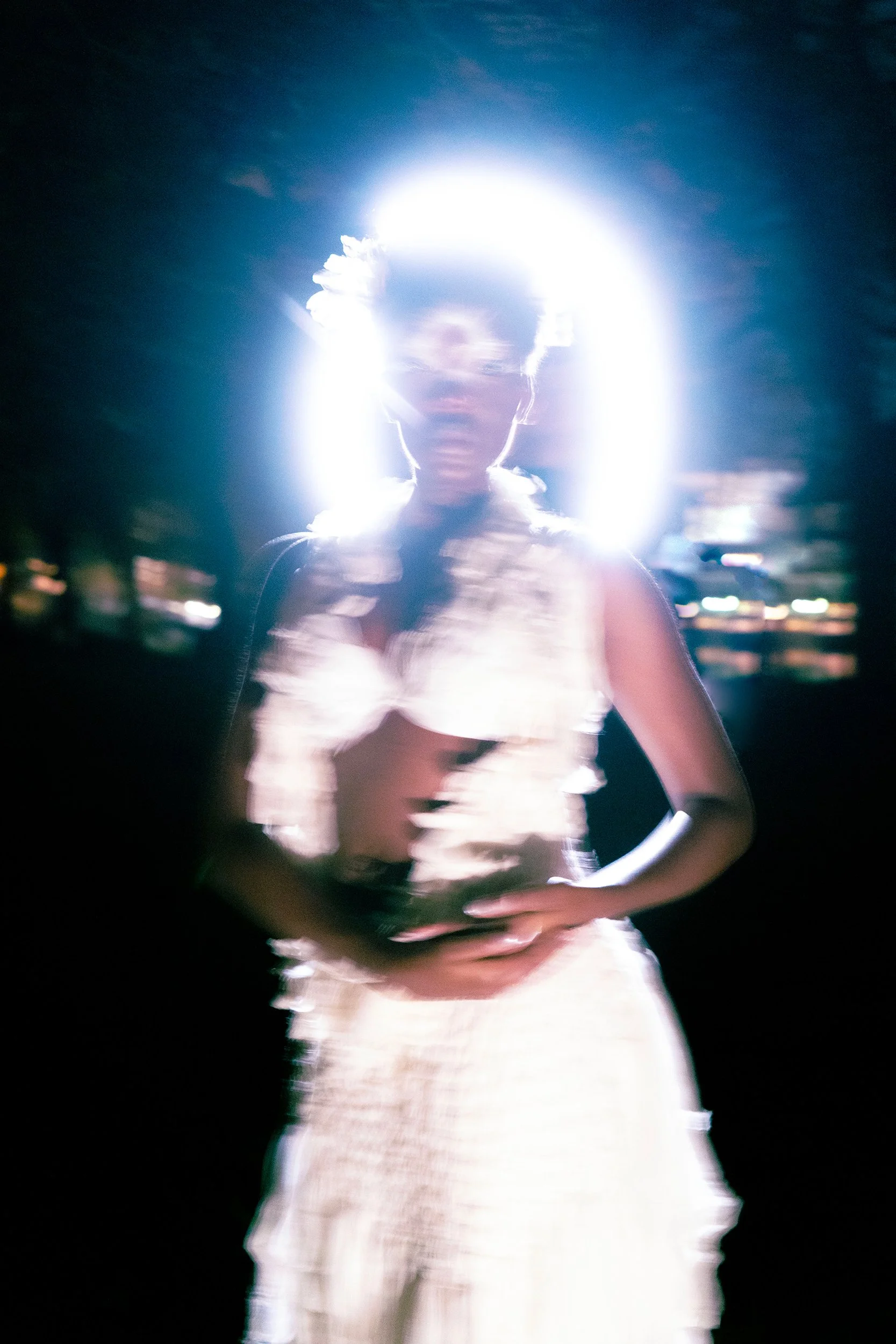 A woman standing outdoors at night, illuminated by a bright light behind her, creating a glow around her head. She is wearing a sleeveless dress and has her hands resting on her waist.