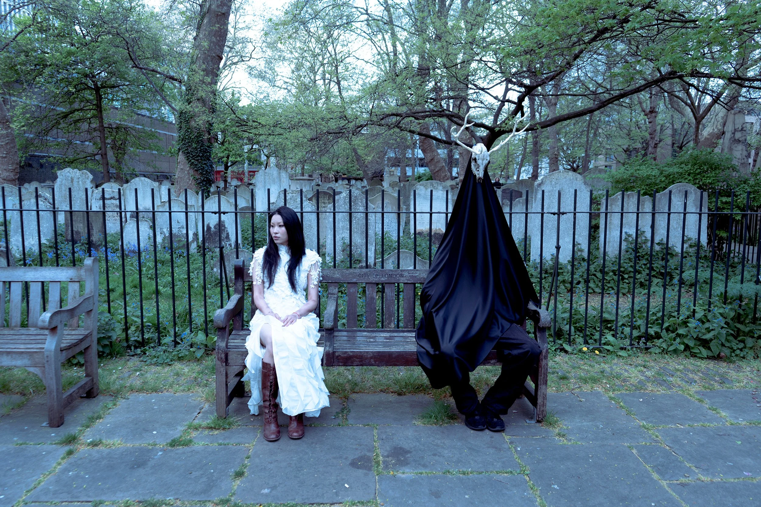A woman in a white dress and brown boots sitting on a park bench. A person in black with a deer skull on their head and black cape is standing next to her. The setting is a green park with trees and a fence, and old gravestones in the background.
