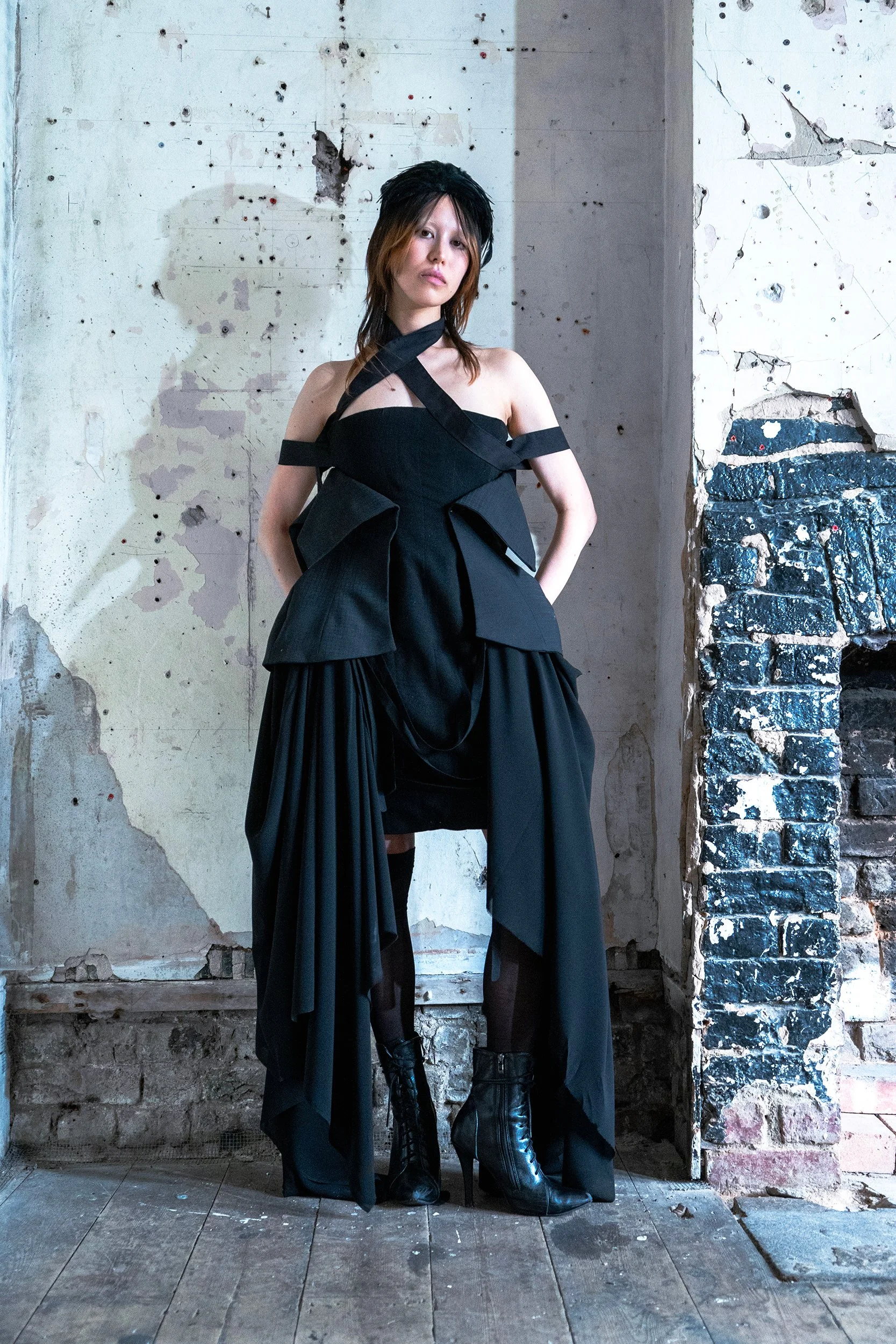 A woman wearing a black off-shoulder dress with crisscross straps, lace-up boots, and black stockings standing against a distressed wall with peeling paint and cracks.