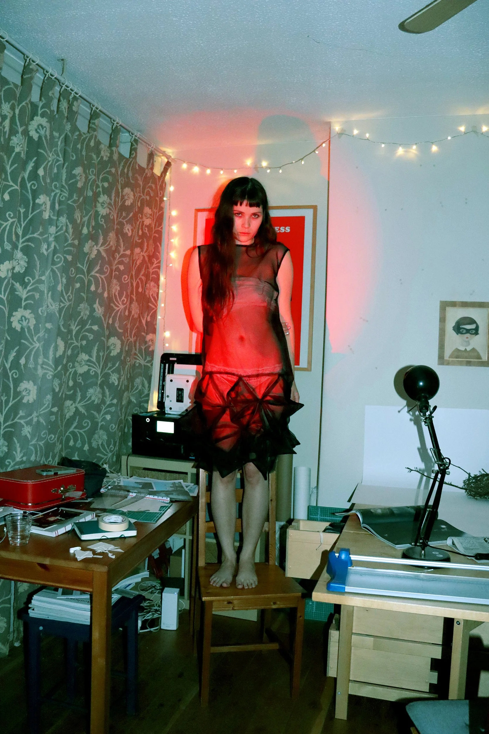 Woman standing on a chair wearing a sheer red dress in a cluttered room with various objects and fairy lights on the wall.