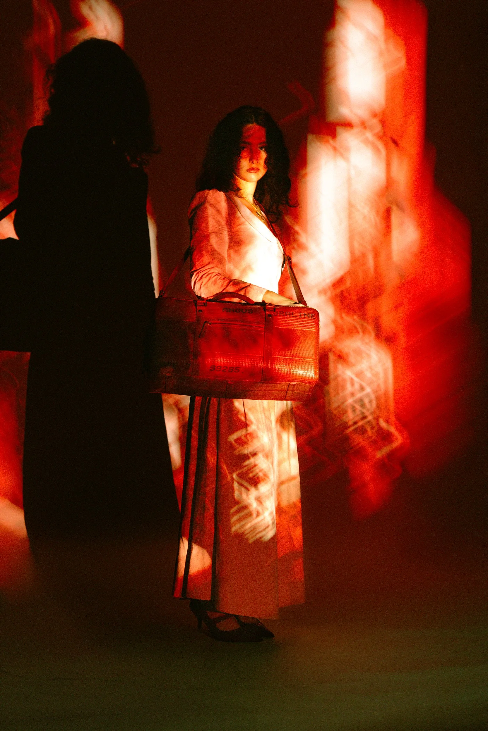 A woman standing on stage with dramatic red lighting, wearing a long, flowing dress and black heels, holding a large red bag, with a shadowed figure partially visible on the left and red textured background.