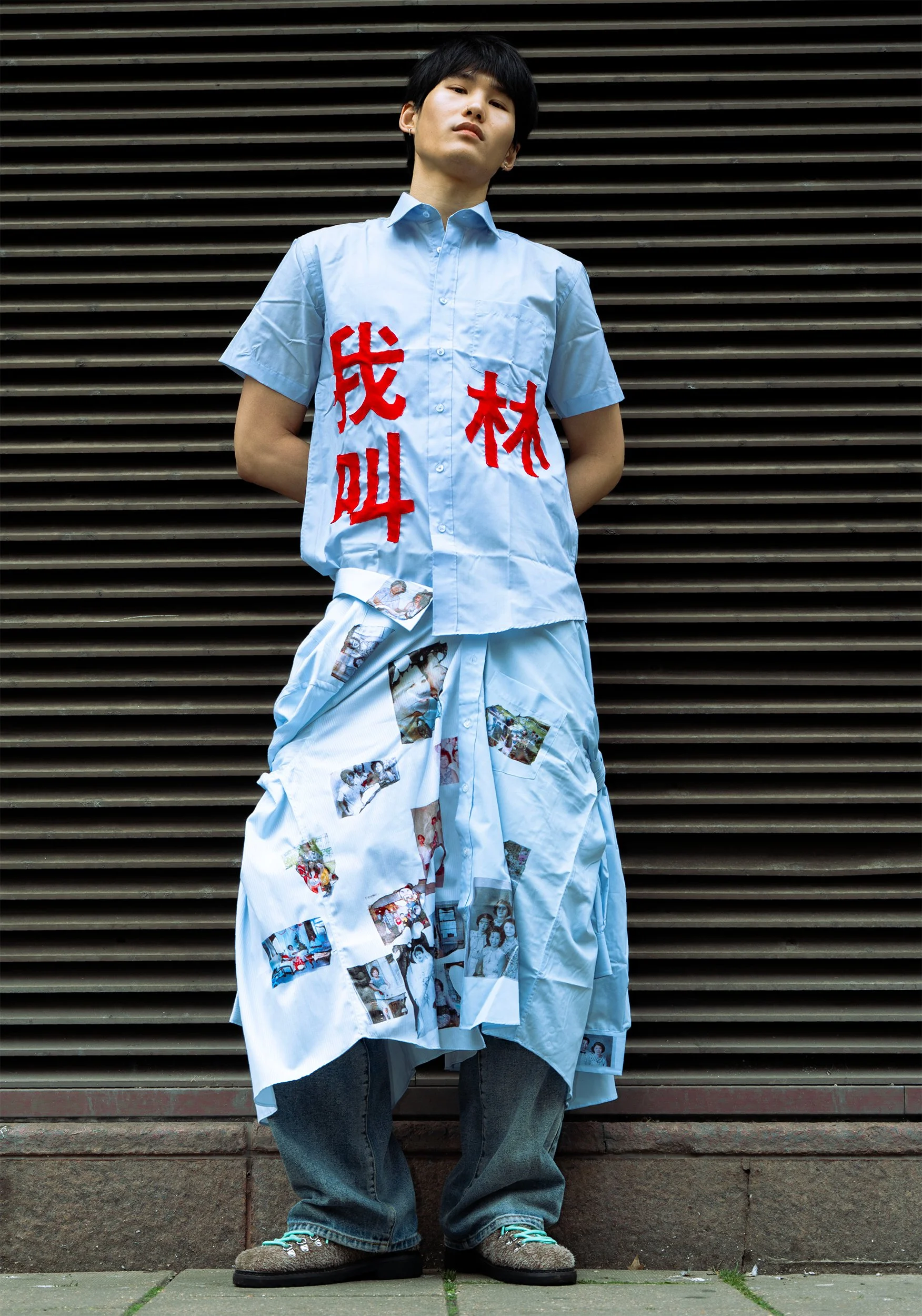 A person standing against a horizontal slatted metal wall, wearing a light blue button-up shirt with red Chinese characters and photographs printed on it, and baggy blue jeans. Their hands are behind their back, and they are looking slightly downward with a serious expression.