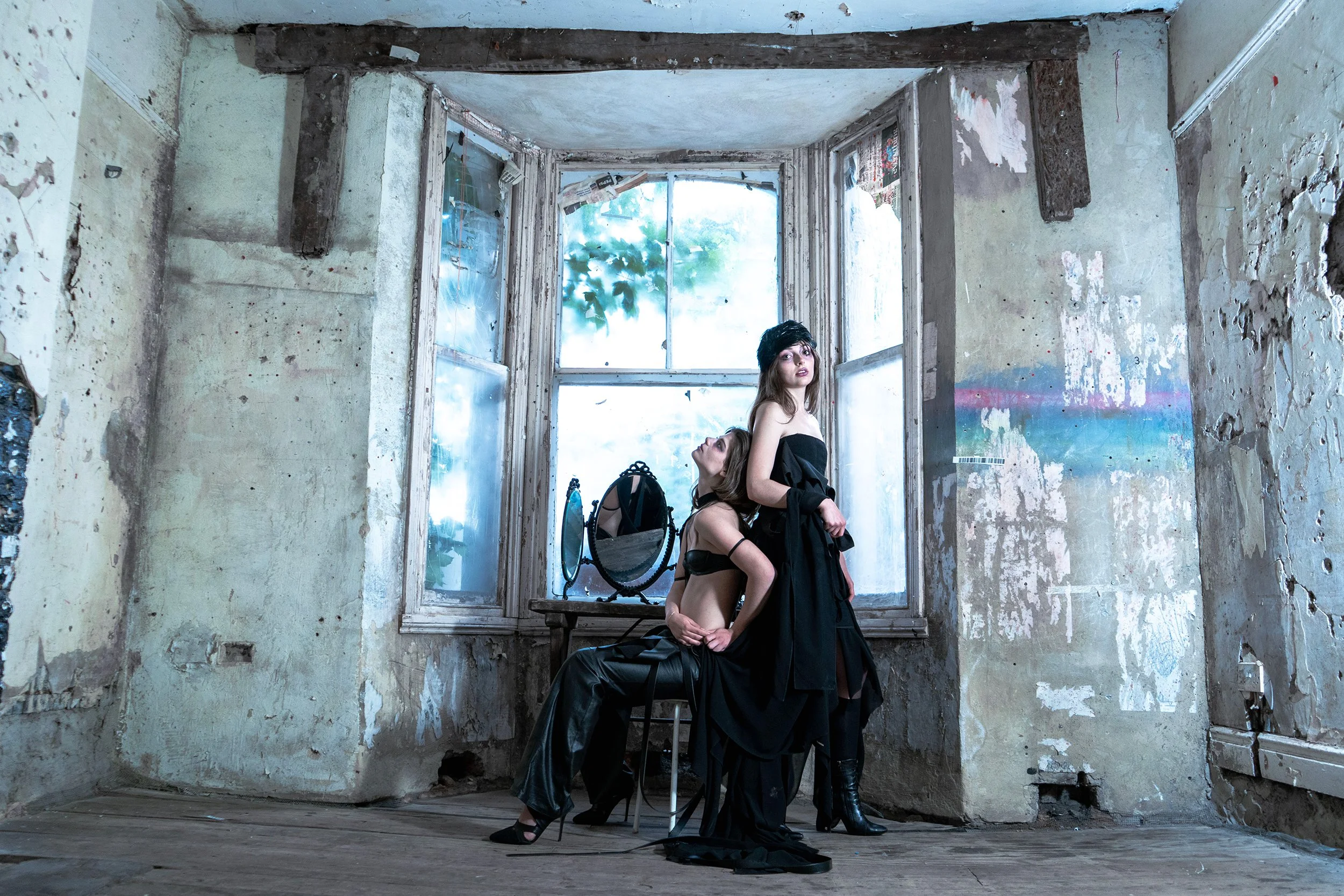 Two women in black clothing and high heels pose in an abandoned, dilapidated room by a window, with peeling paint on the walls and a mirror on a chair between them.